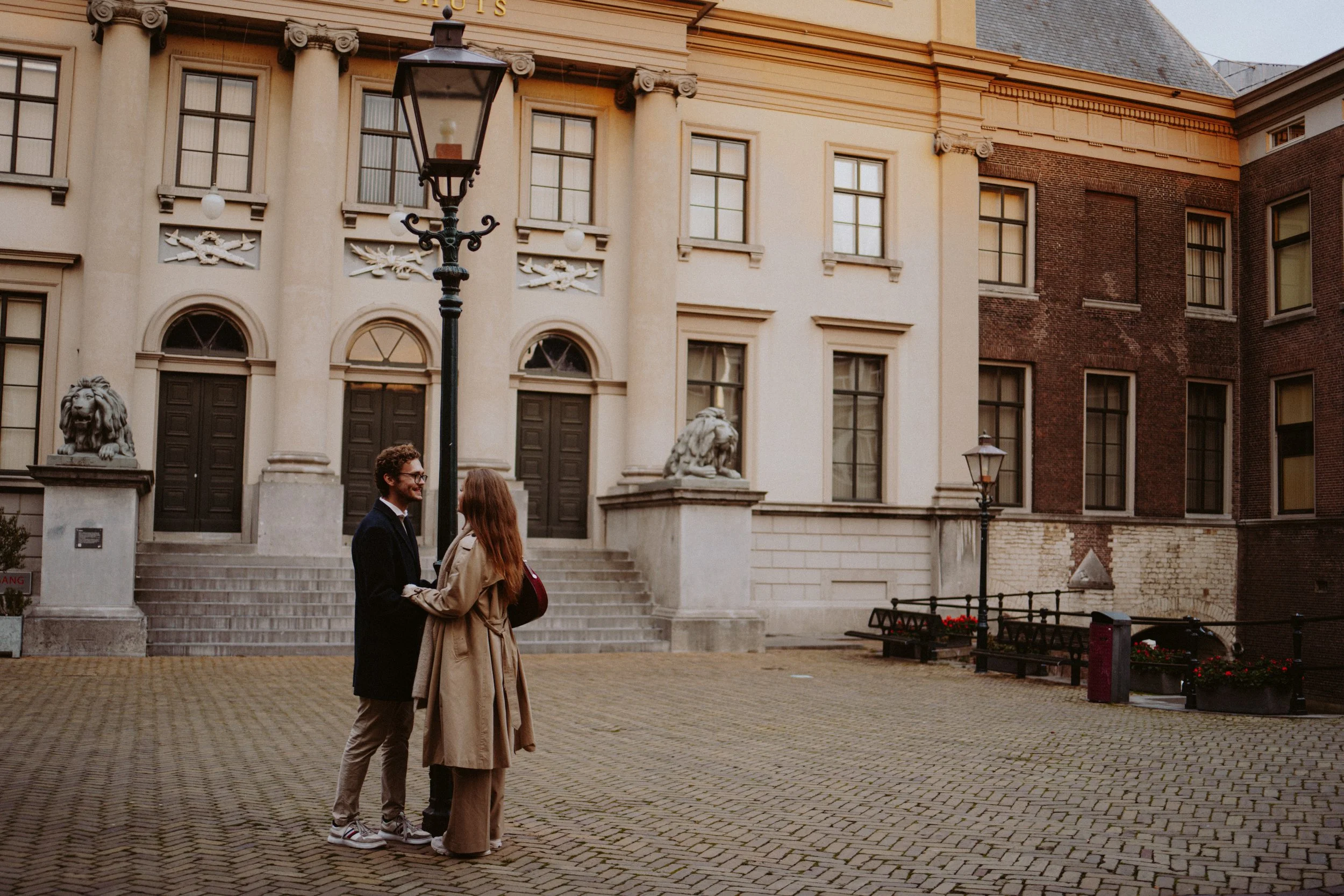 A man and woman standing close together and talking on a brick plaza in front of a large, ornate building with columns, statues of lions on the stairs, and lampposts, during daytime.