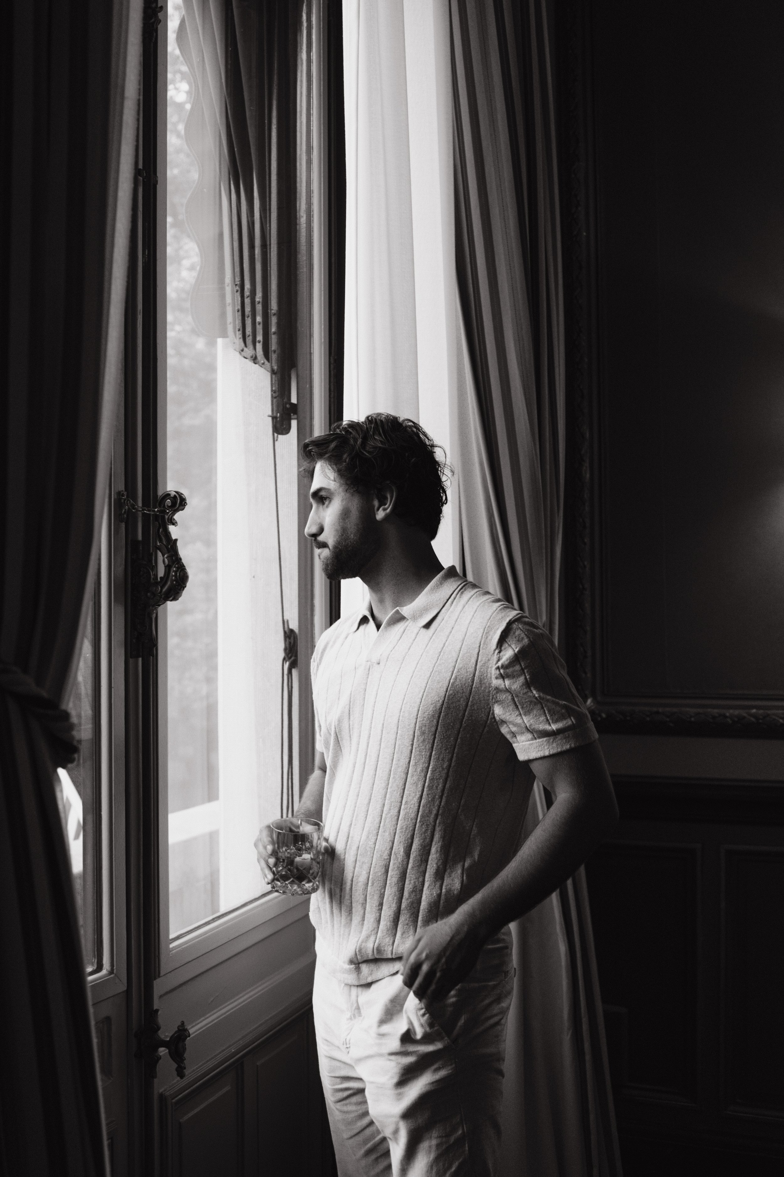 A man with dark, wavy hair and a beard standing indoors by a window, holding a glass, looking out, wearing a striped short-sleeve shirt.