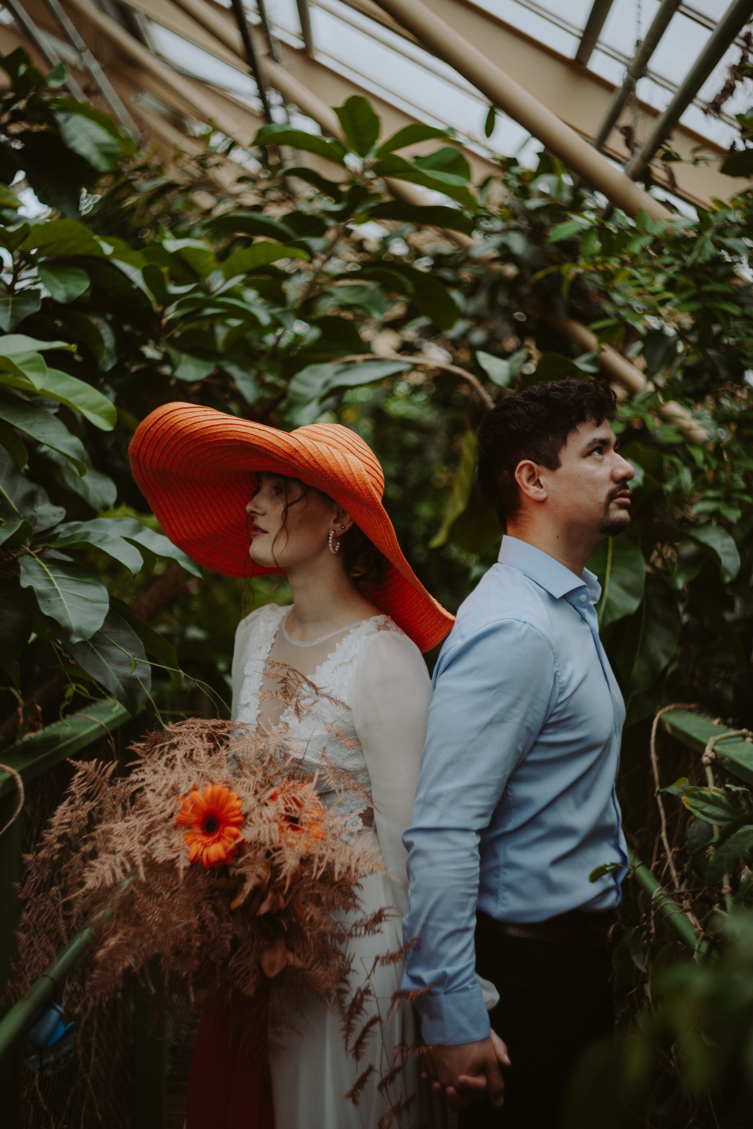 A woman wearing a large orange sun hat and white dress, and a man in a light blue shirt, holding hands and standing back-to-back in a greenhouse filled with green plants and flowers.