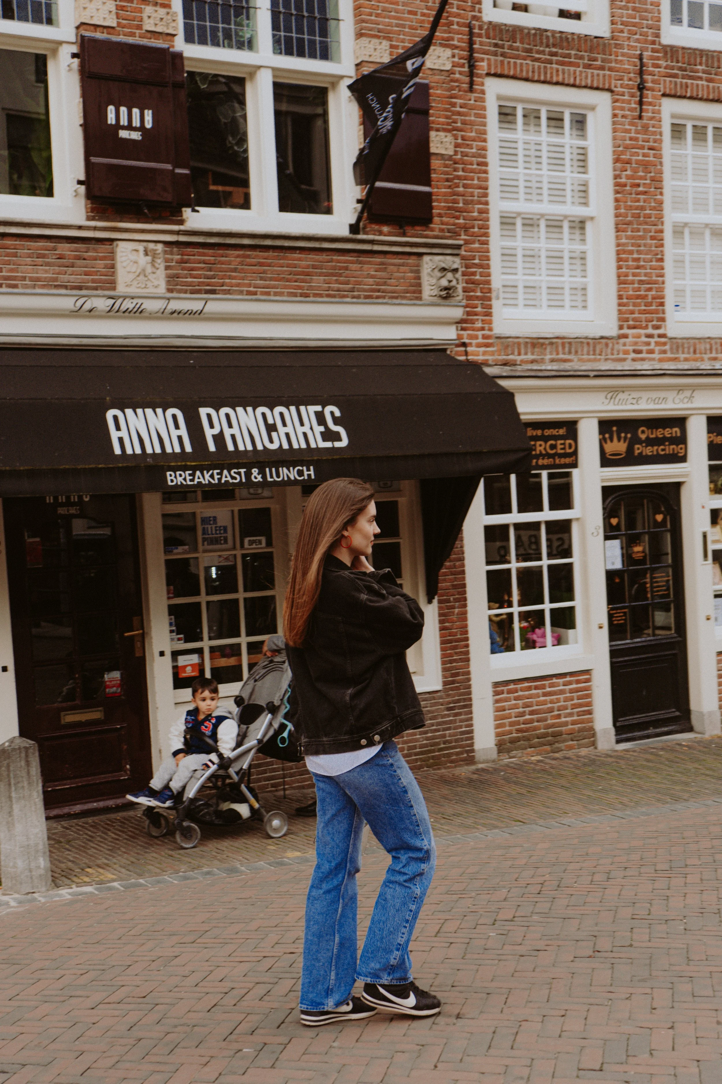 A woman walking on a brick sidewalk pushing a stroller with a young boy sitting inside. The scene is in front of a cafe with black awnings labeled 'Anna Pancakes' and informational signs. The woman has long brown hair, is dressed in a black jacket, b