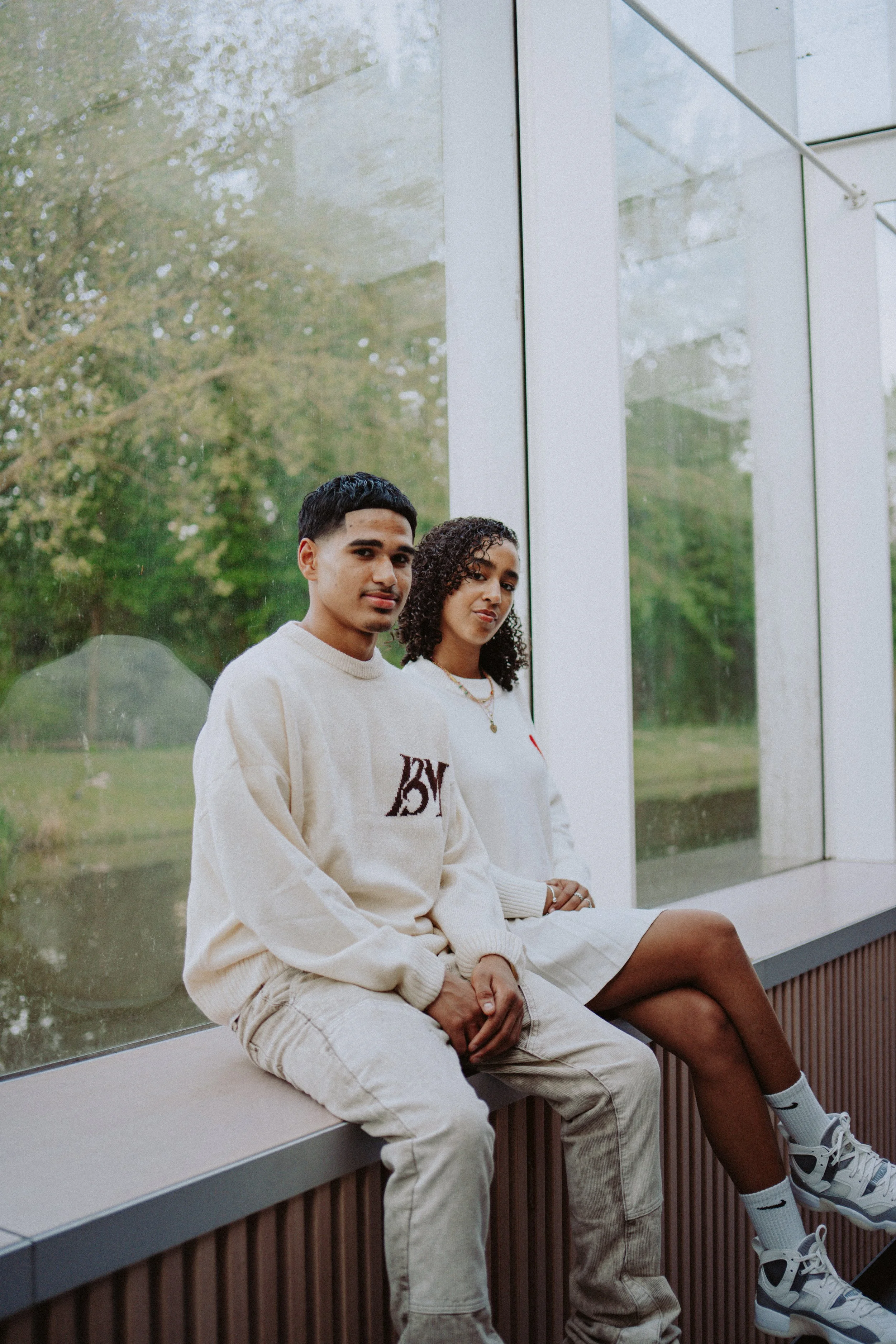 Two young adults sitting on a bench near a large window with trees visible outside, both wearing casual, neutral-colored clothing.