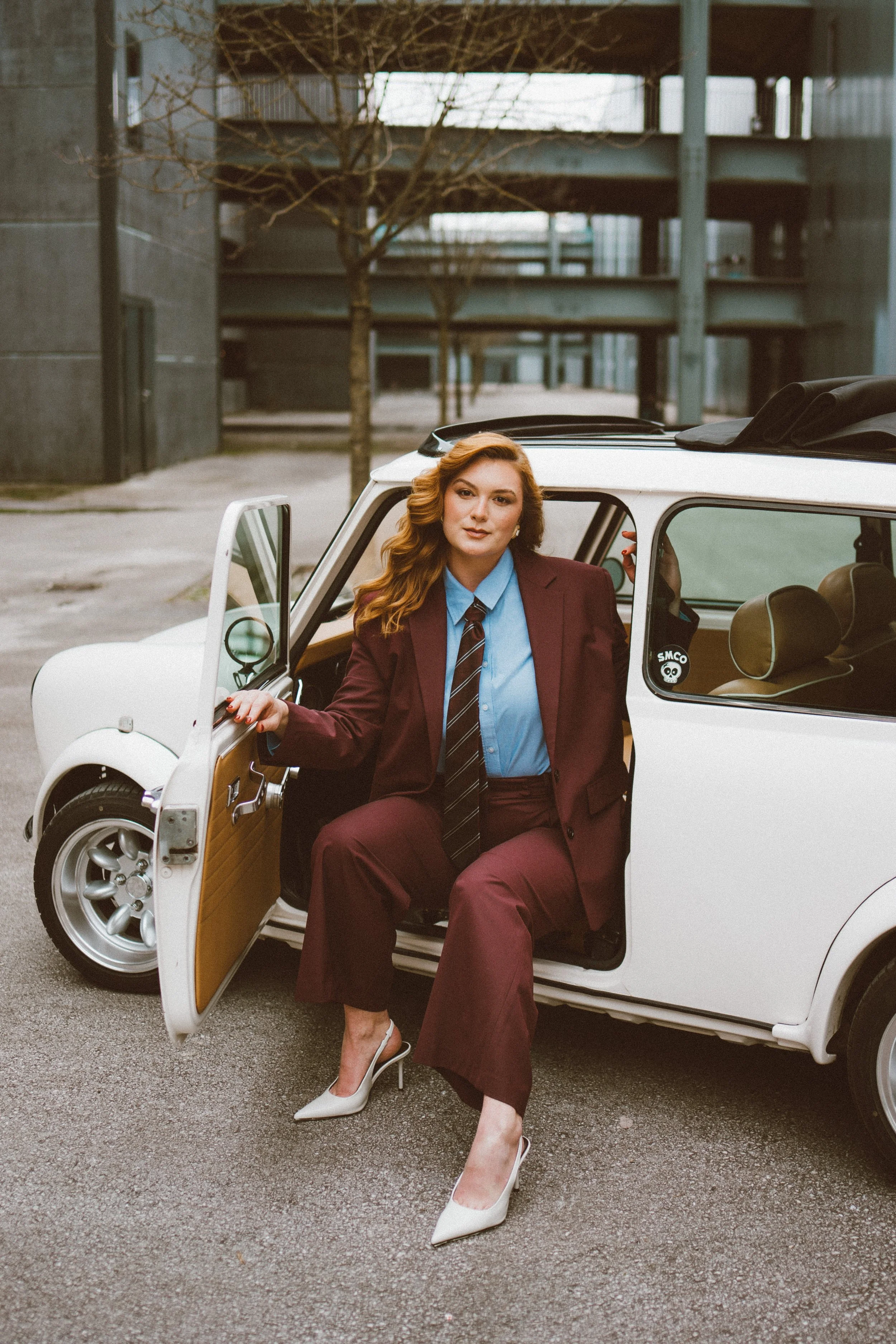 A woman dressed in a maroon suit, light blue shirt, and a striped tie is sitting in a vintage white car with the door open. She has long, wavy red hair and is wearing white high heels. The background includes a modern urban setting with concrete stru