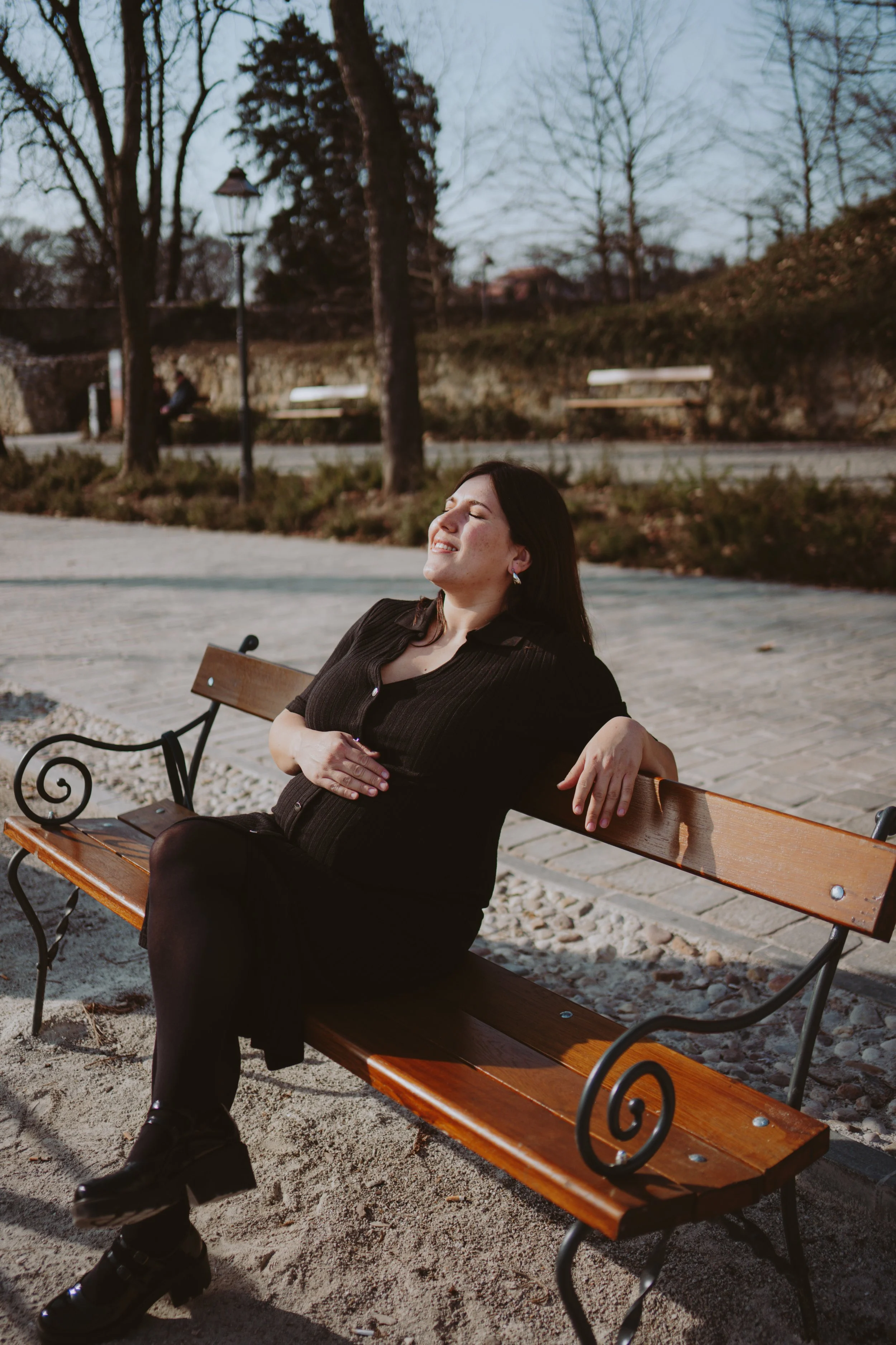 A woman is sitting on a wooden park bench with black metal armrests and legs, relaxing with her eyes closed and a smile on her face. She is wearing a black outfit and earrings. The park has leafless trees, bushes, a stone wall, a lamppost, and anothe