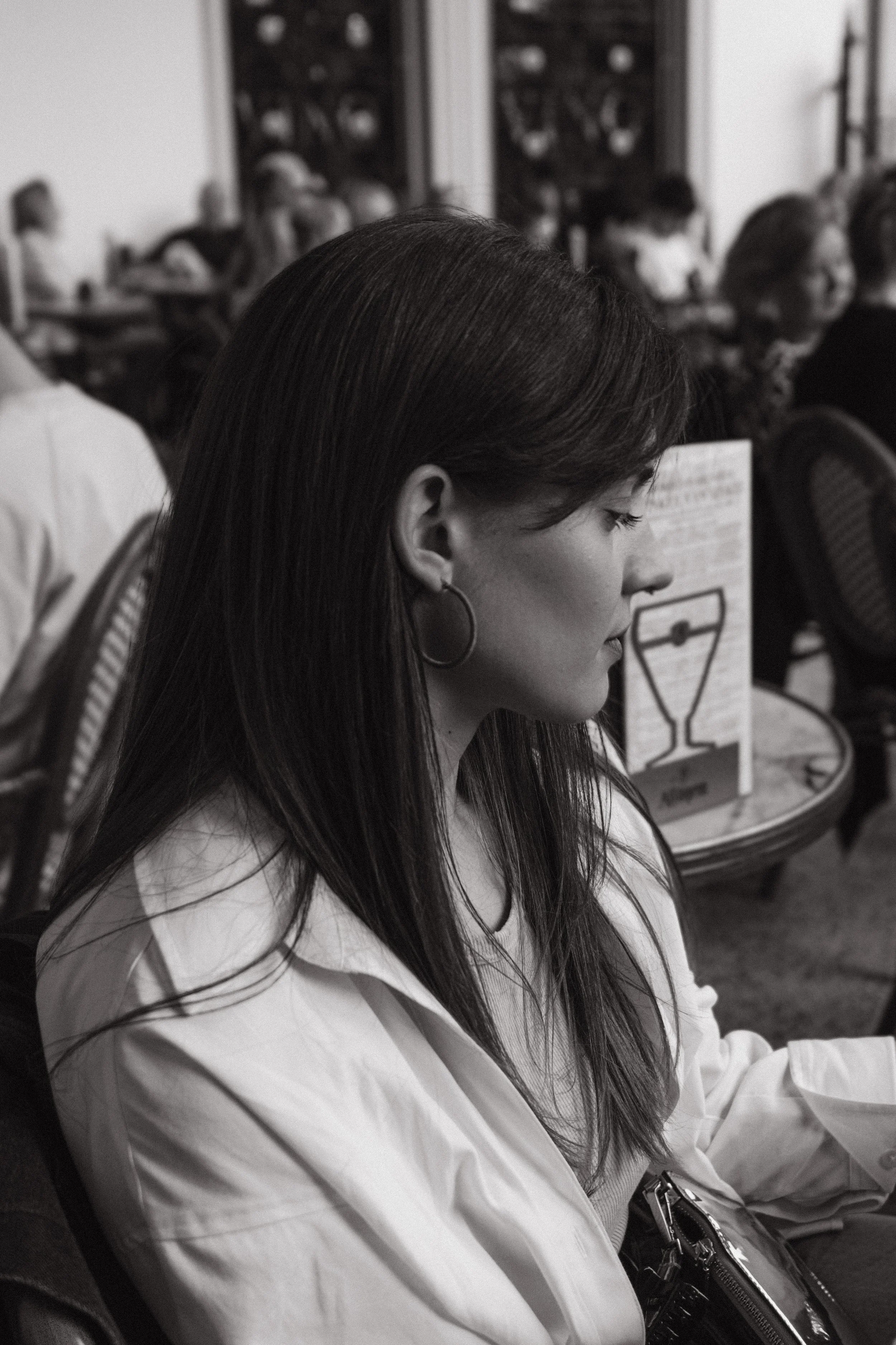 A woman with long dark hair and hoop earrings sitting with her eyes closed, wearing a white shirt, in a room with other people and chairs, possibly in a classroom or meeting setting.