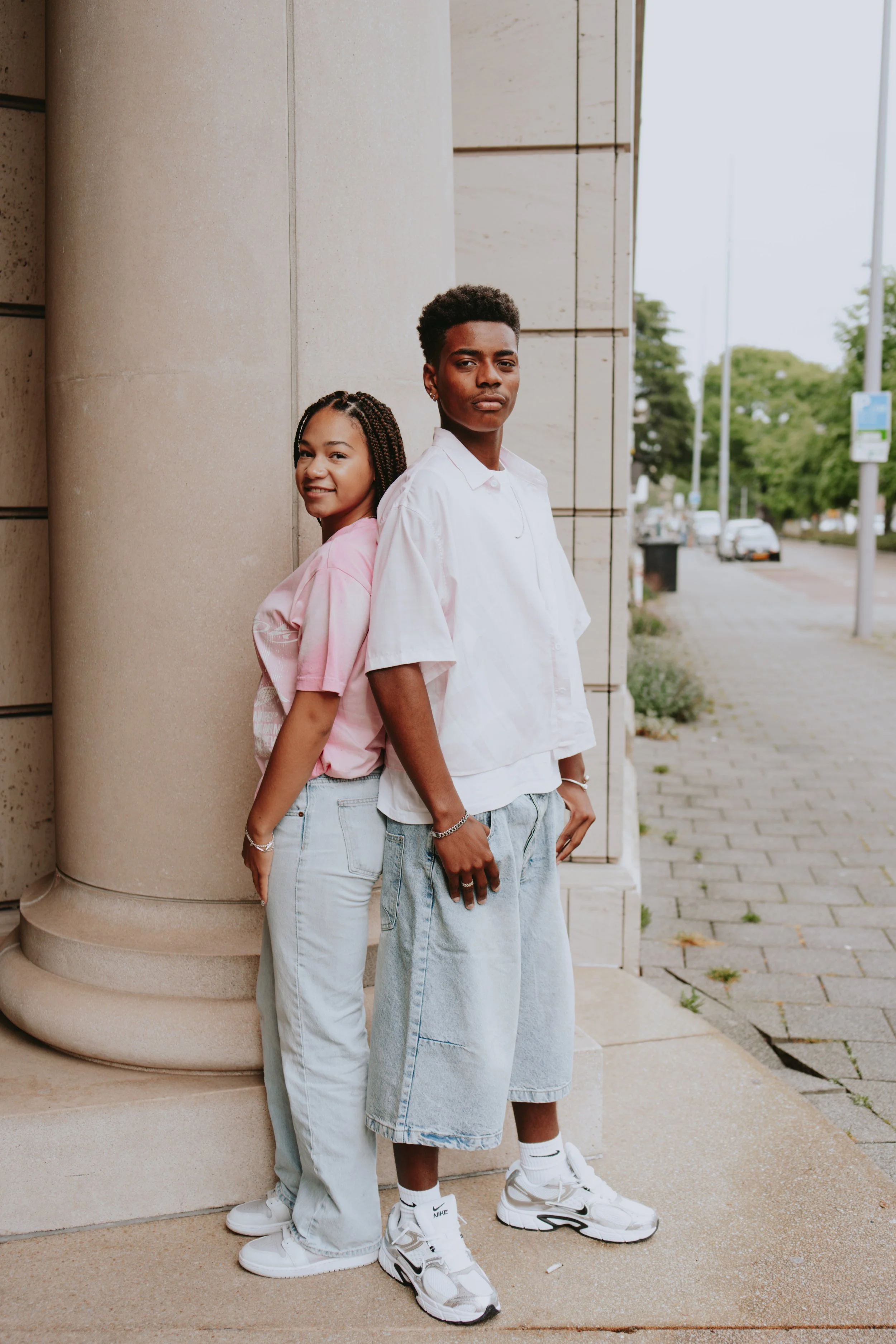 Two young people standing back-to-back outdoors next to a large stone column, with a city sidewalk and trees in the background.