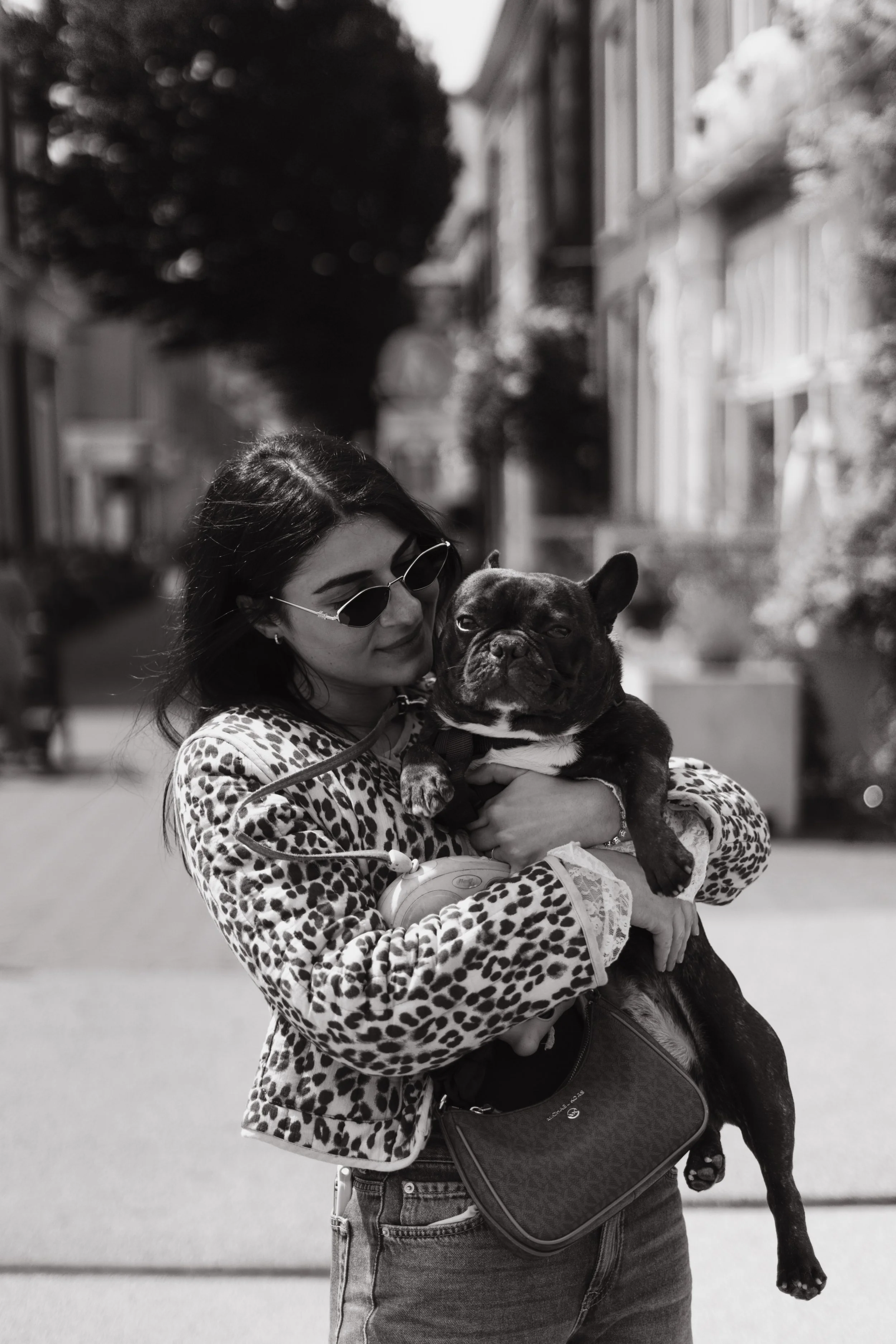 A woman wearing sunglasses and a leopard print jacket holding a French Bulldog in her arms on a city street.