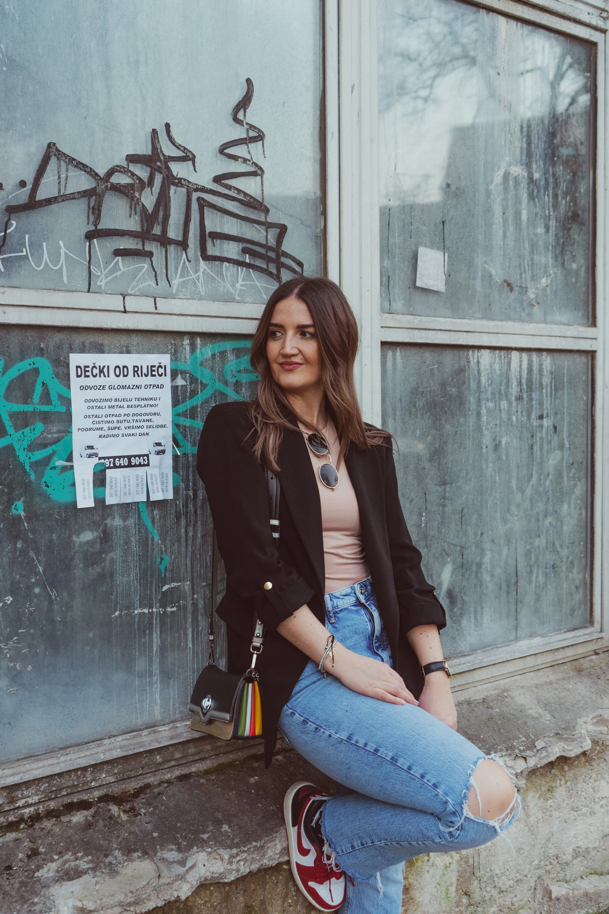 A young woman sitting outdoors against a weathered glass wall with graffiti. She has long brown hair, wears a black blazer, beige top, ripped blue jeans, and red sneakers. She carries a black bag with colorful striped details and wears sunglasses aro