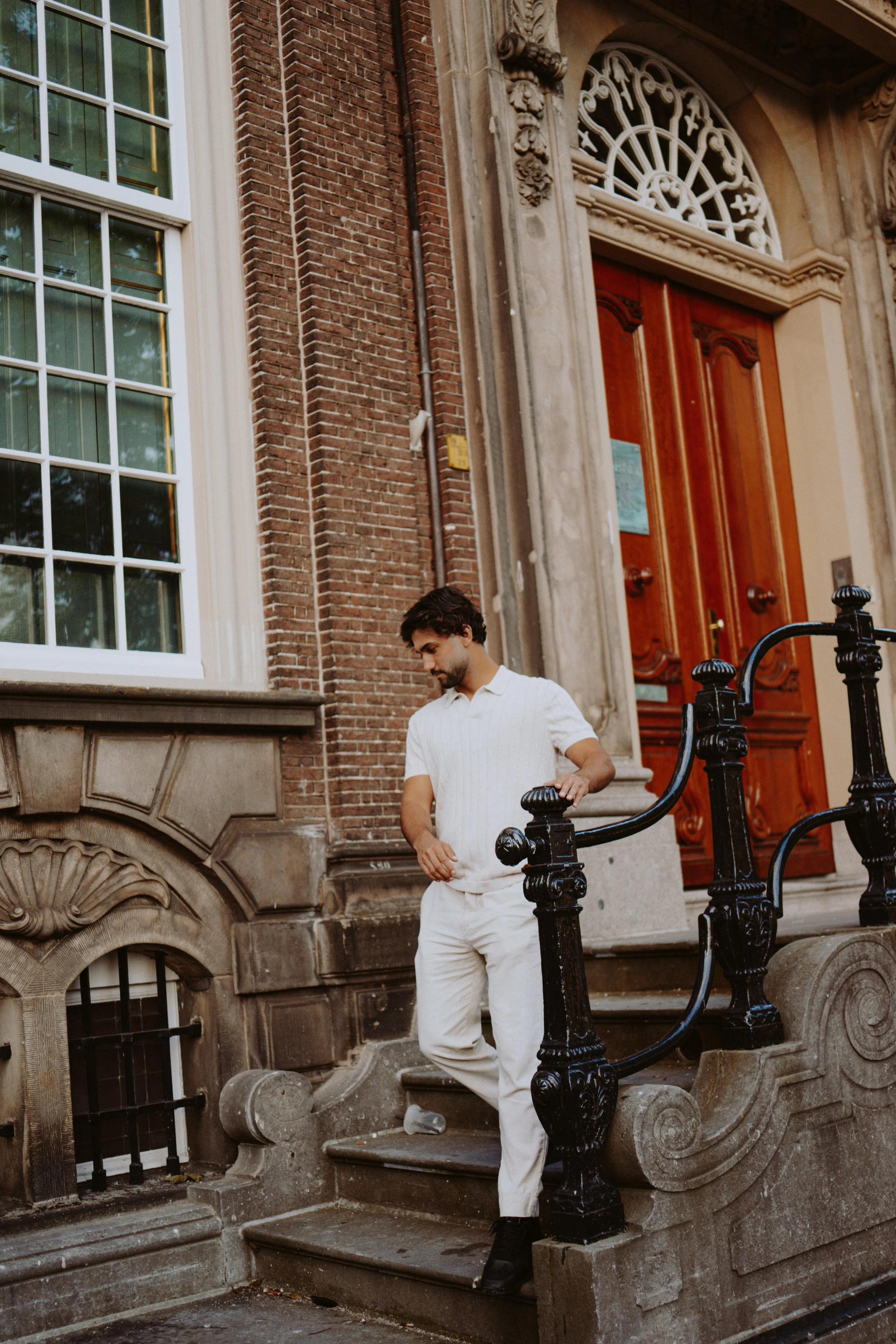 A man with dark hair and a beard, dressed in a white shirt and light-colored pants, standing on the steps outside a historic brick building with ornate stone detailing and a large wooden door. He is looking down at the steps.