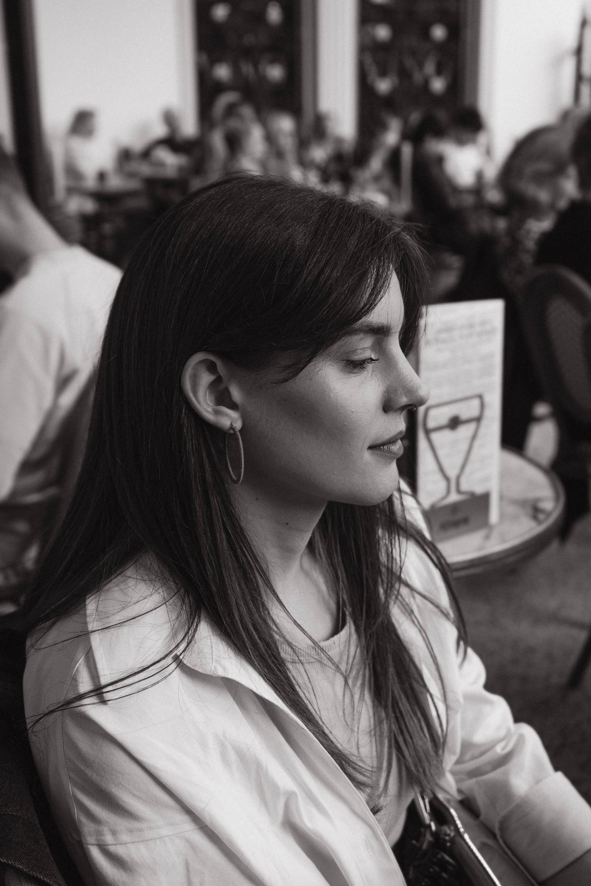 A young woman with long dark hair, wearing a light-colored top and hoop earrings, is seated with her eyes closed, in a room filled with people in the background.