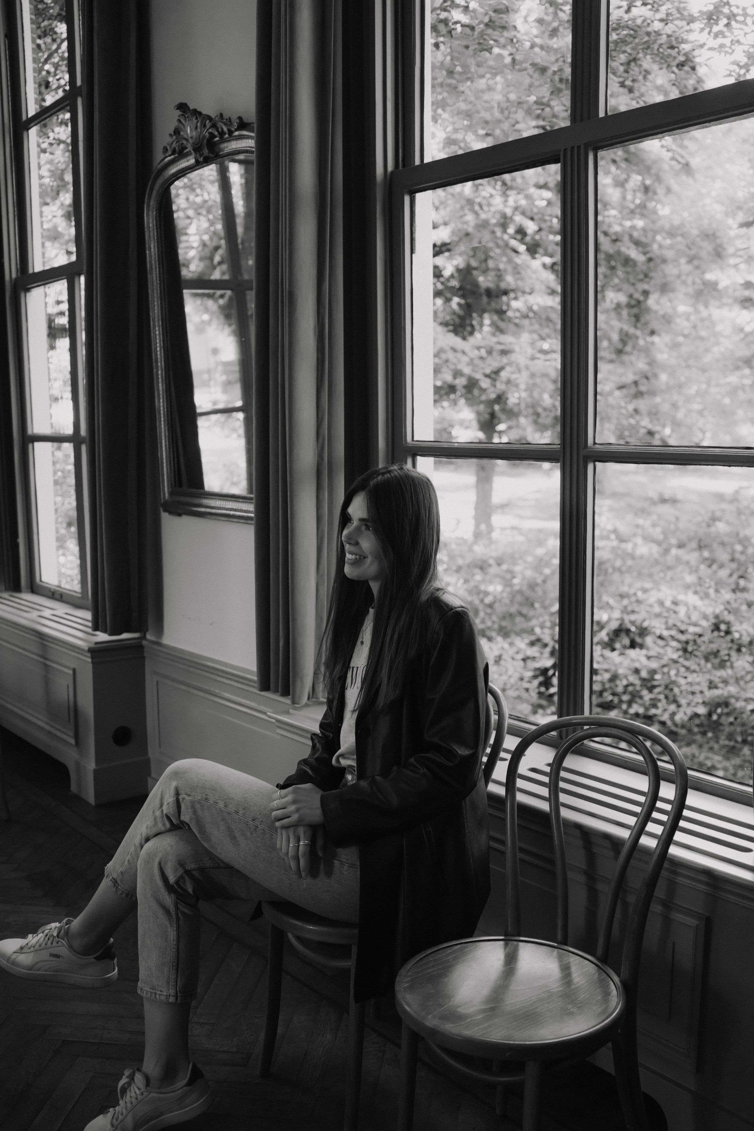 A woman with long hair sitting on a chair in front of large windows with a scenic outdoor view.