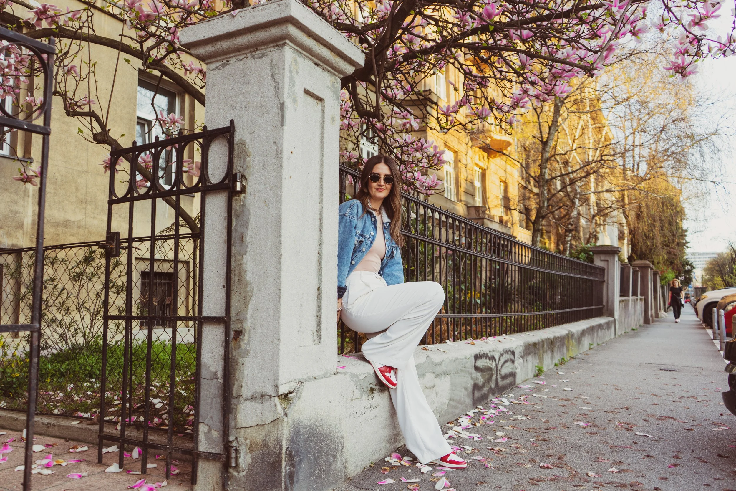A young woman wearing sunglasses, a denim jacket, and white pants is sitting on a low stone wall next to a black metal fence. She is amidst blooming pink magnolia flowers and pink flower petals scattered on the sidewalk. There are building facades an