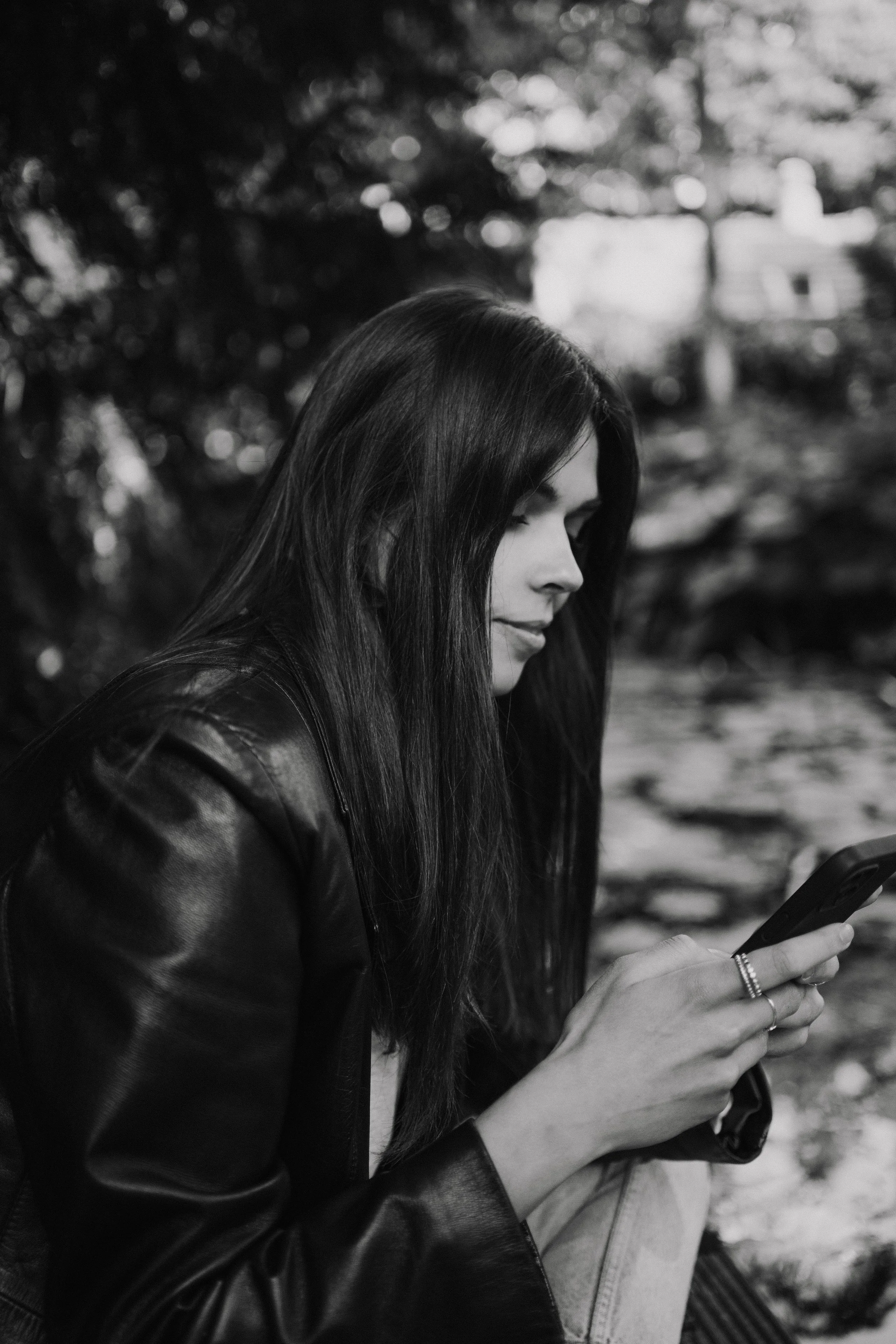 A woman with long dark hair, wearing a leather jacket, looking at her phone, with a blurred background of trees and houses.