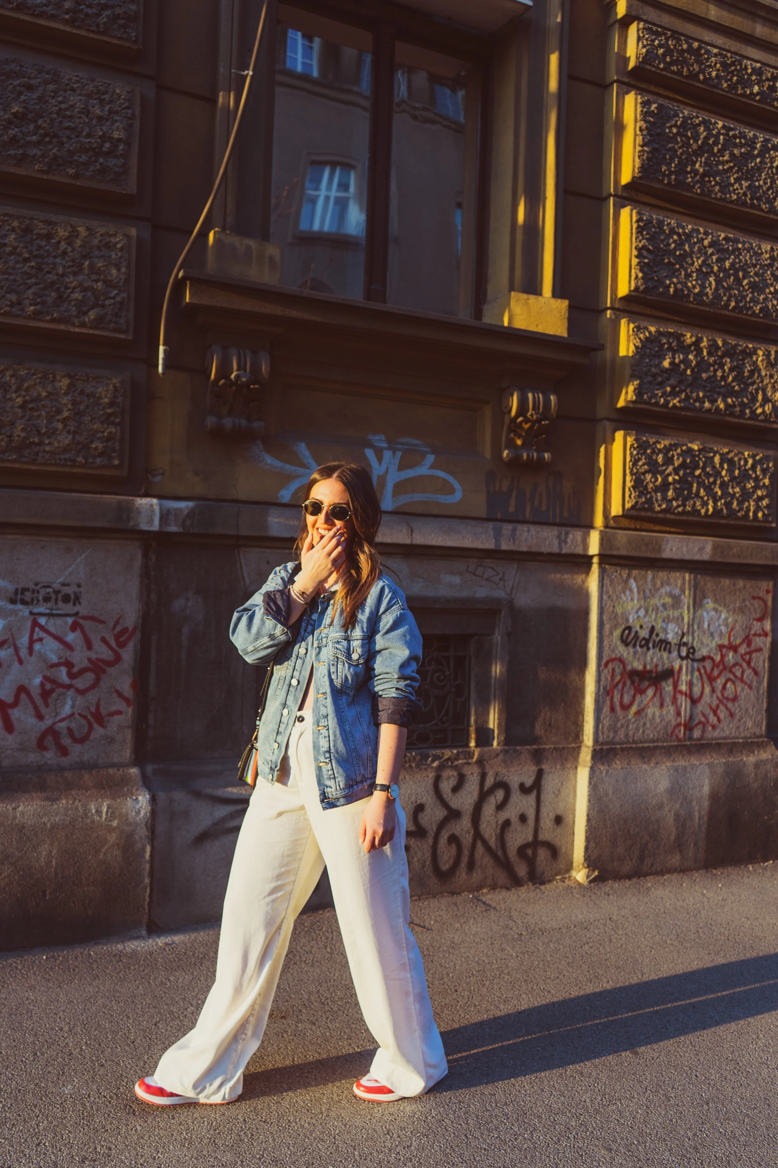 A woman with long hair wearing sunglasses, a denim jacket, white pants, and red and white sneakers, smiling and covering her mouth with her hand, standing on a sidewalk in front of a graffiti-covered building with a window above her.