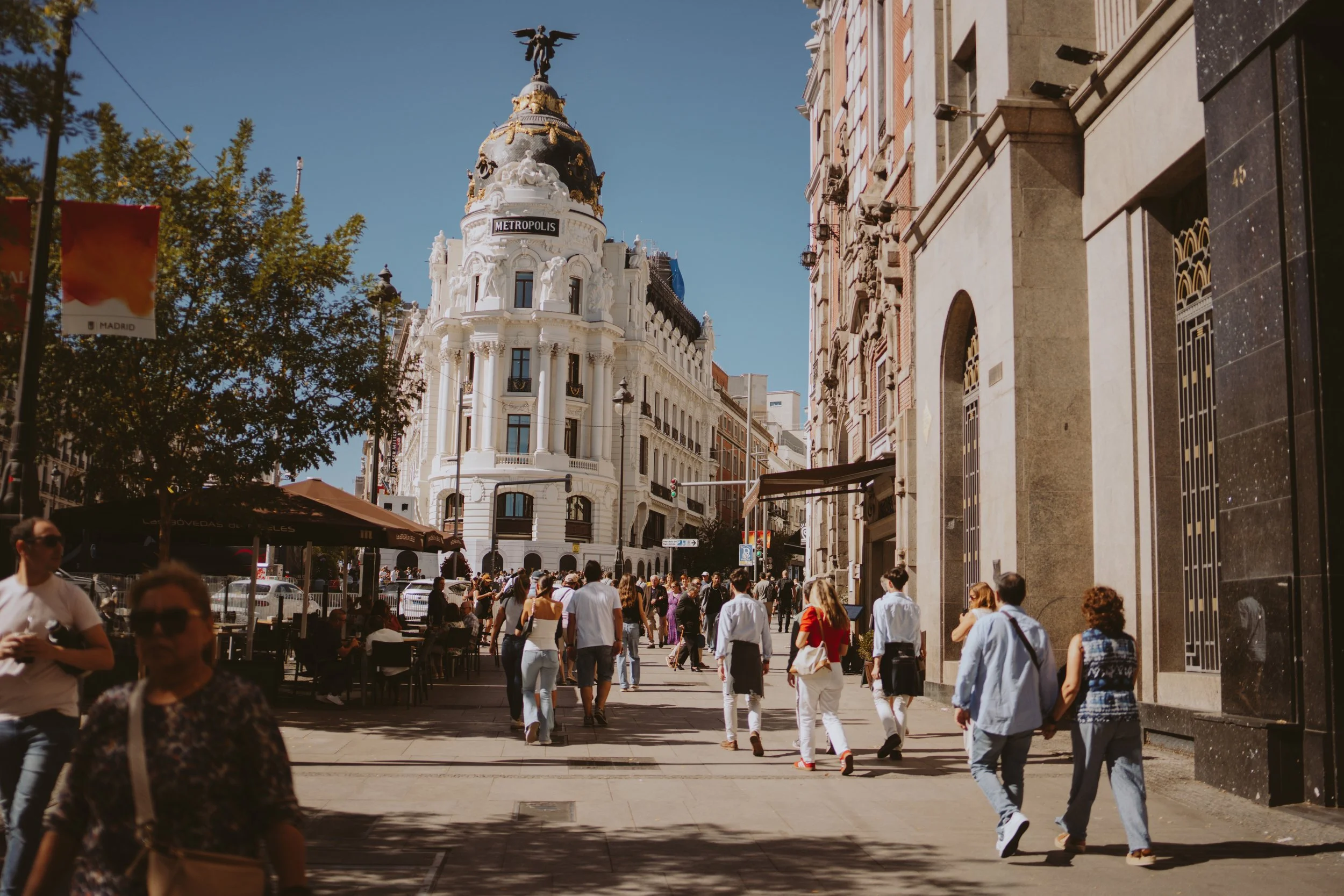 Crowd of people walking along a city street with historic buildings, including the Metropolis building with a statue on top, under a clear blue sky.
