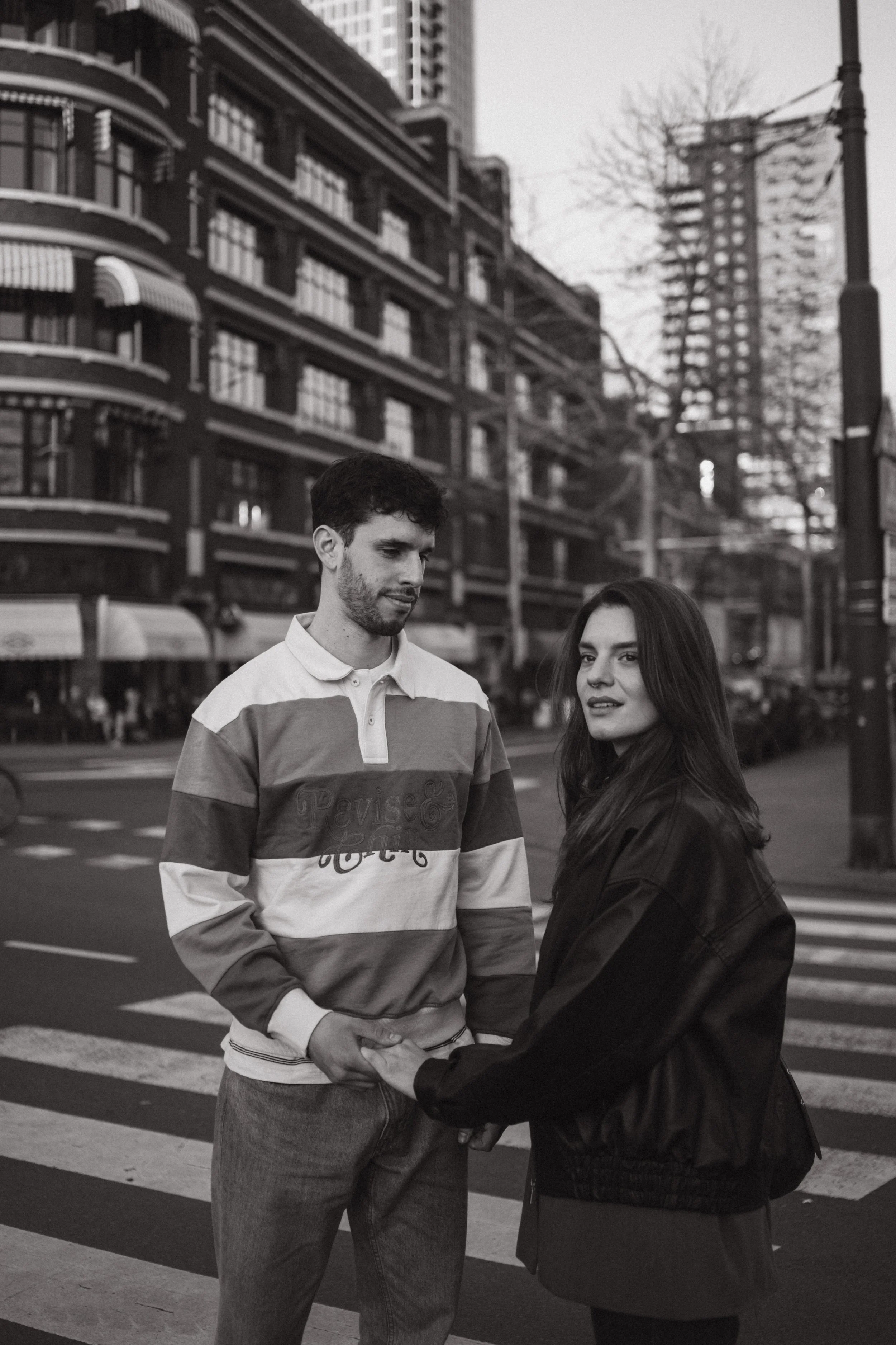A young man and woman holding hands on a city street, with buildings and a tree in the background, in a black and white photograph.