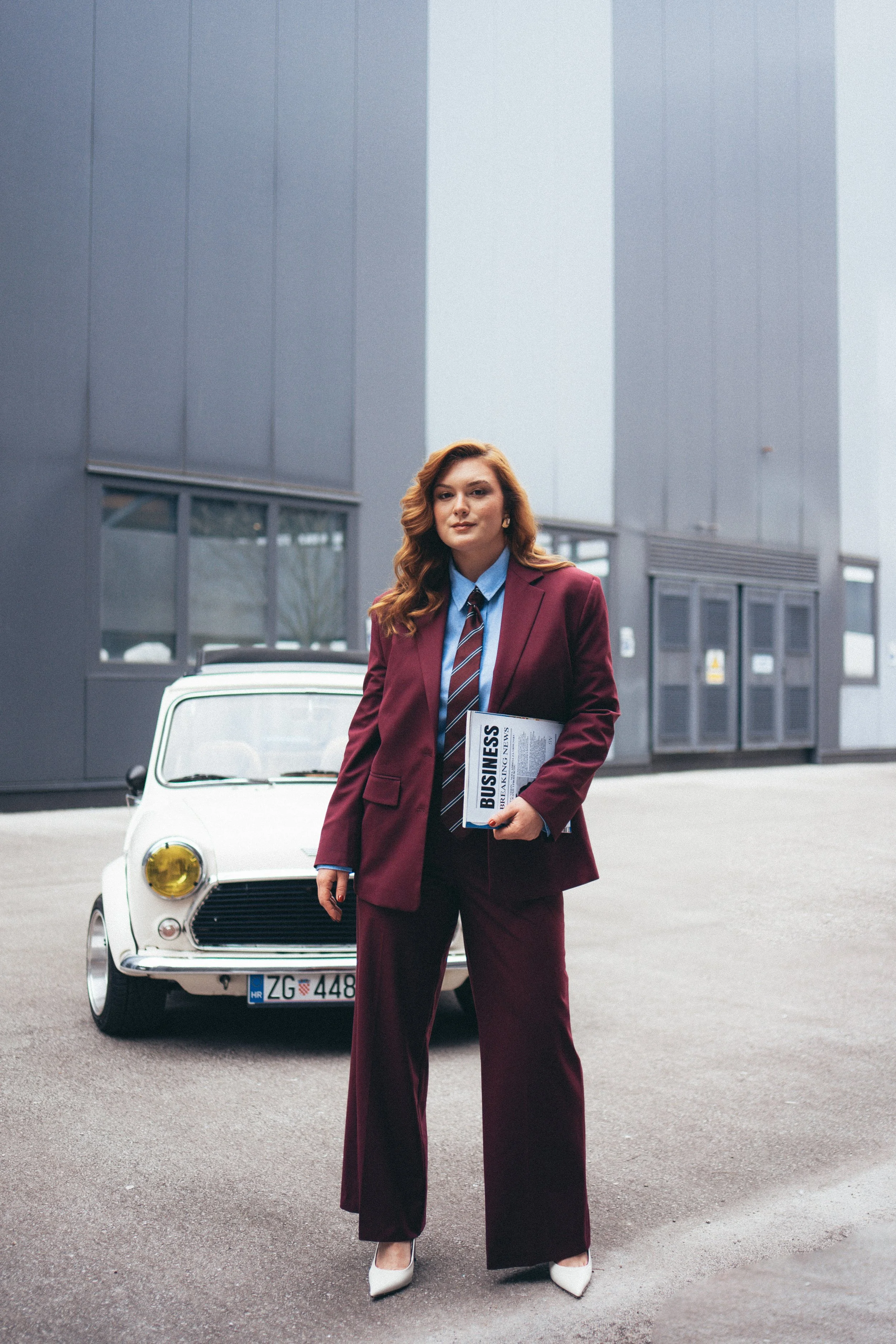 A woman with long red hair in a burgundy suit holding a newspaper labeled 'BUSINESS' stands in front of a white vintage car with yellow headlights, outside a modern building.