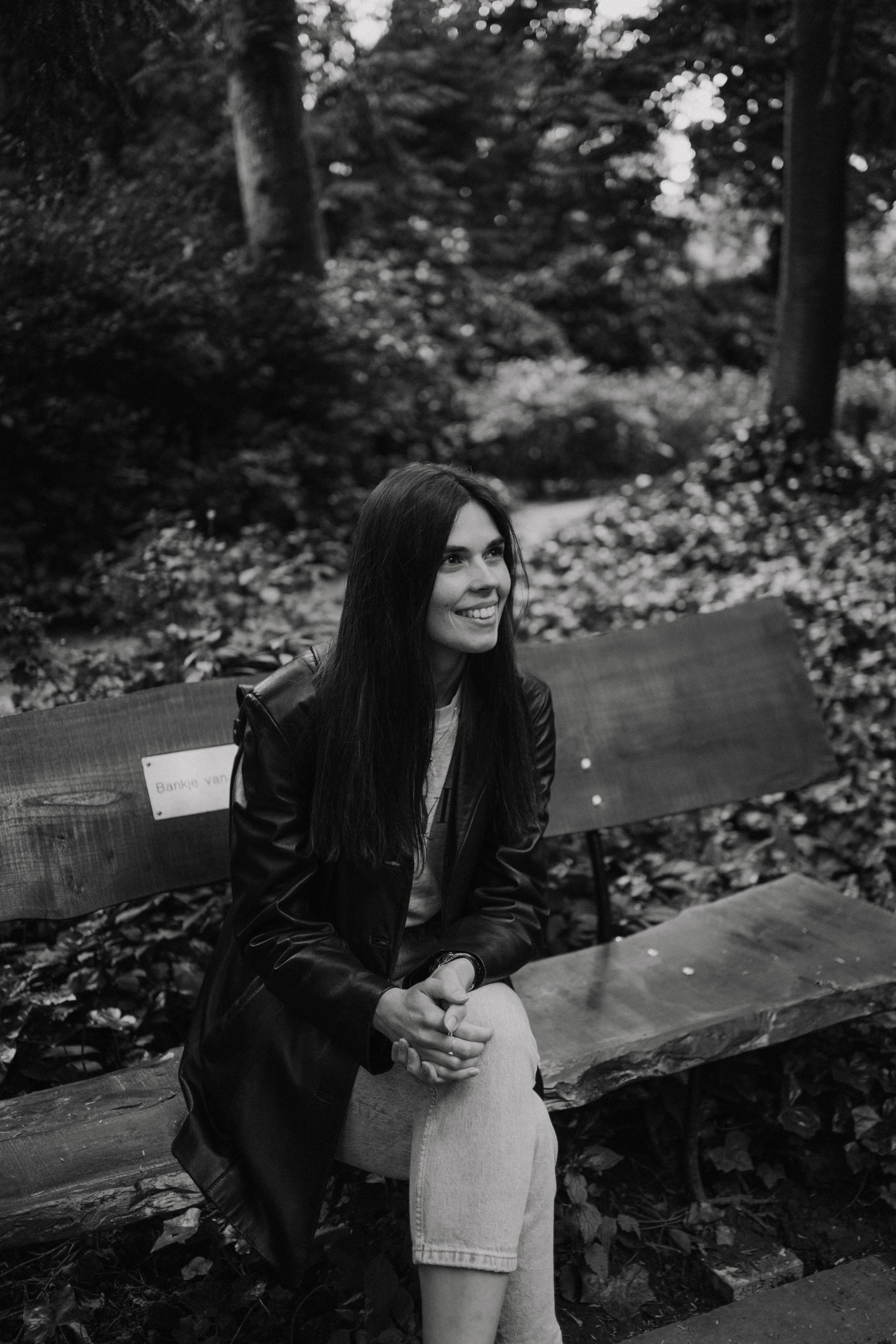 A woman with long dark hair smiling, sitting on a wooden bench in a wooded park, wearing a leather jacket and light shorts.