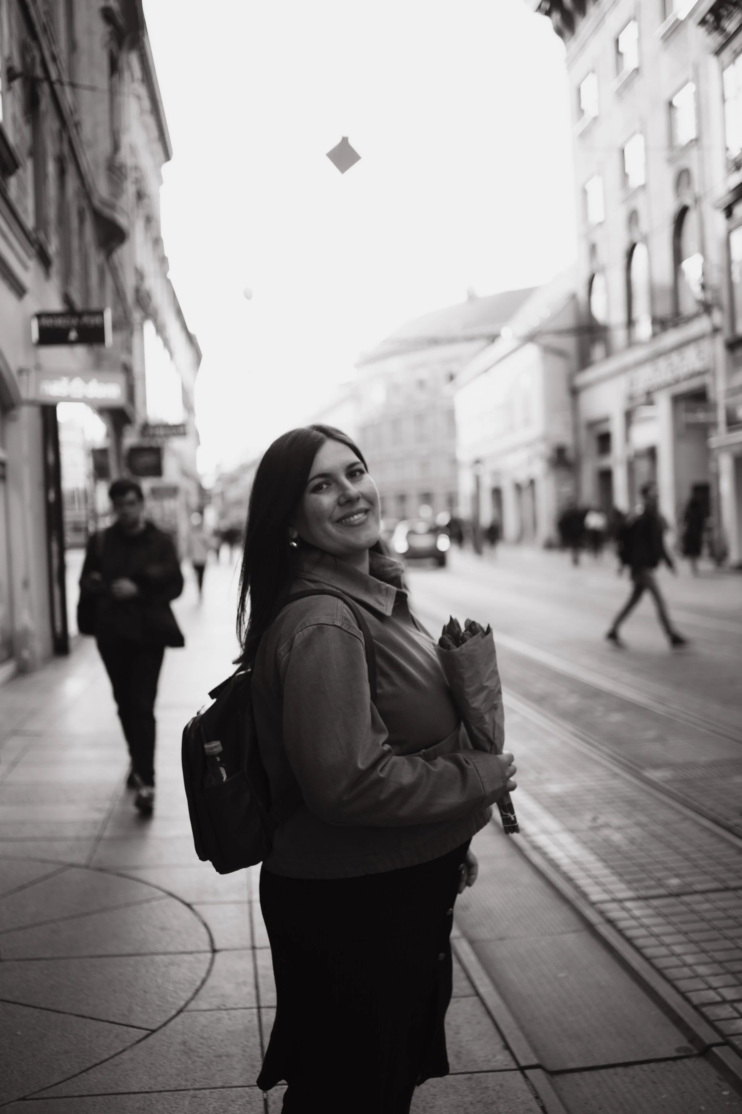 A woman with long dark hair, smiling, standing on a city sidewalk with buildings and other pedestrians in the background, holding a bouquet of flowers.