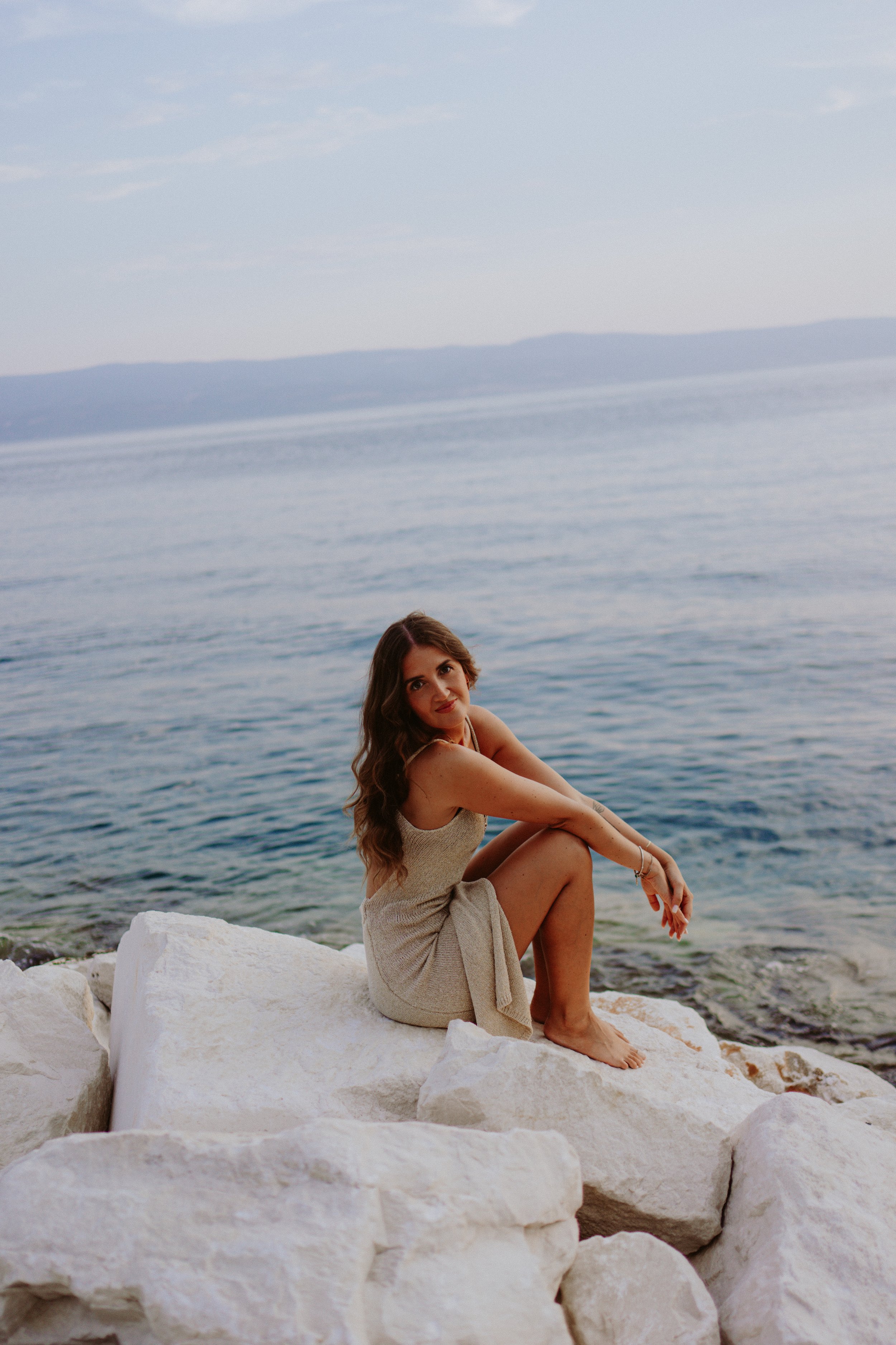 A woman with long brown hair sitting on white rocks by the water, with an ocean and distant mountains in the background, during daylight.
