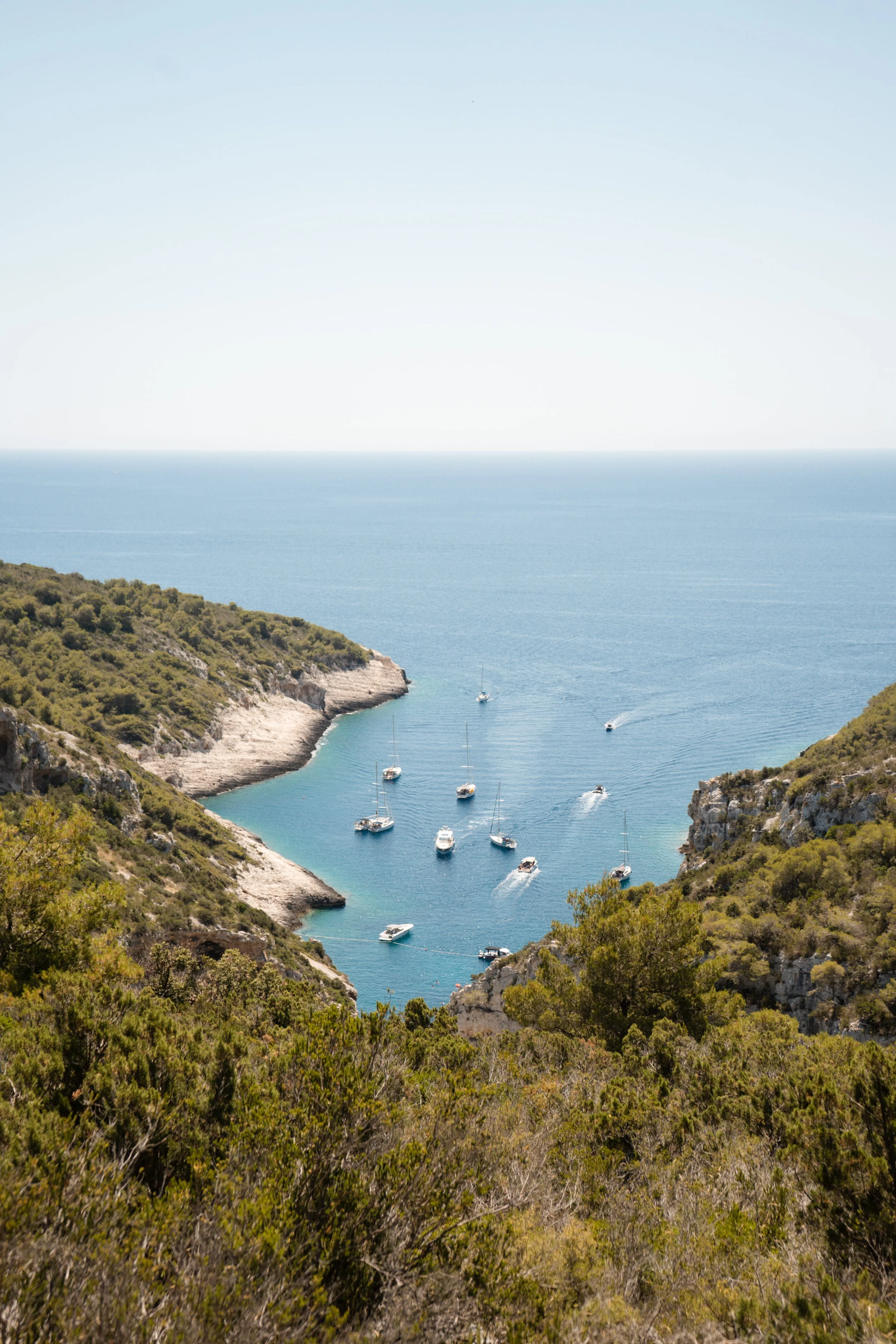 A coastal scene with a narrow waterway between two cliffs covered in green vegetation, leading to a calm bay with several boats anchored and sailing on the blue water under a clear sky.