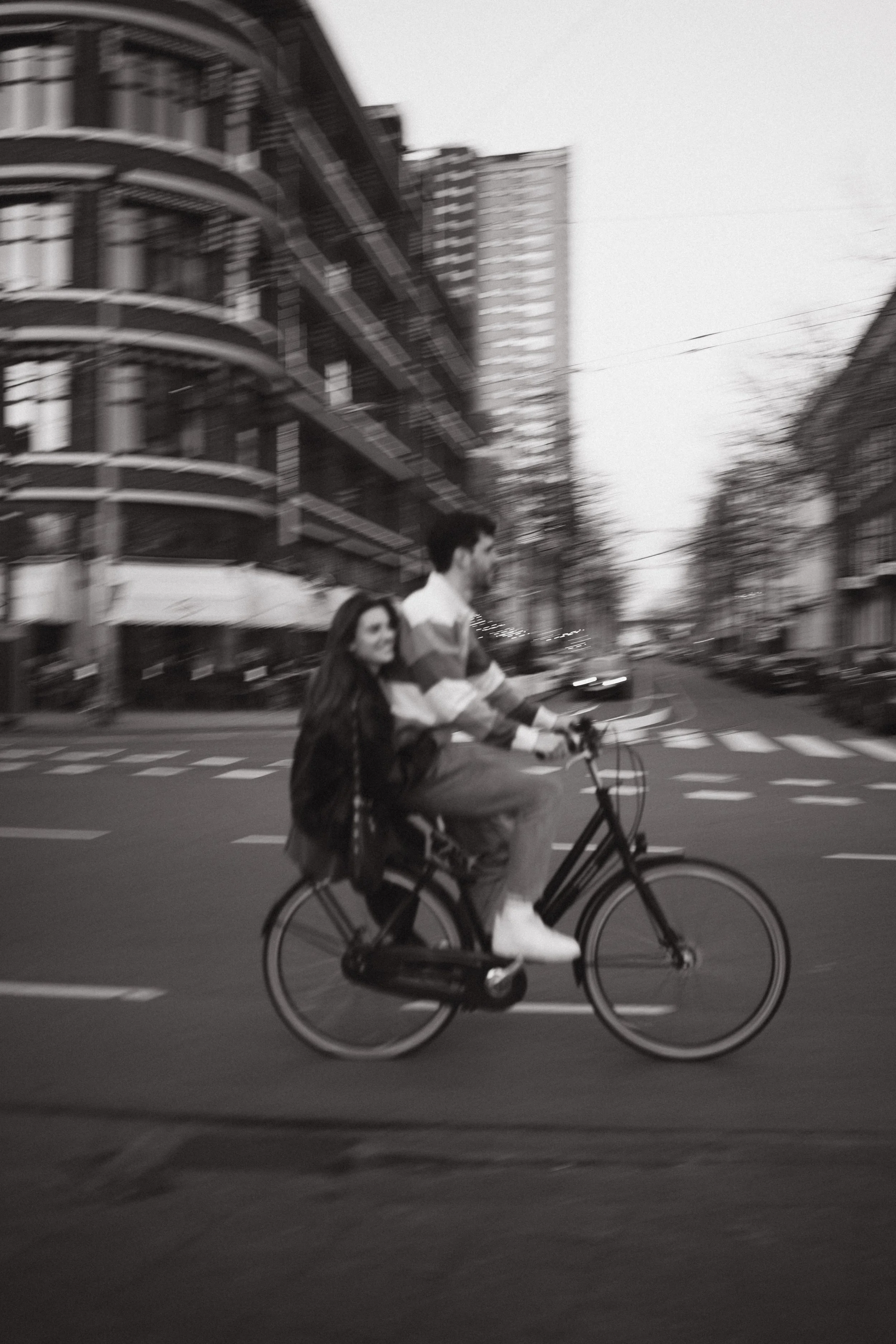 Black and white photo of a man and a woman riding a bicycle together on a city street, with buildings and leafless trees in the background.
