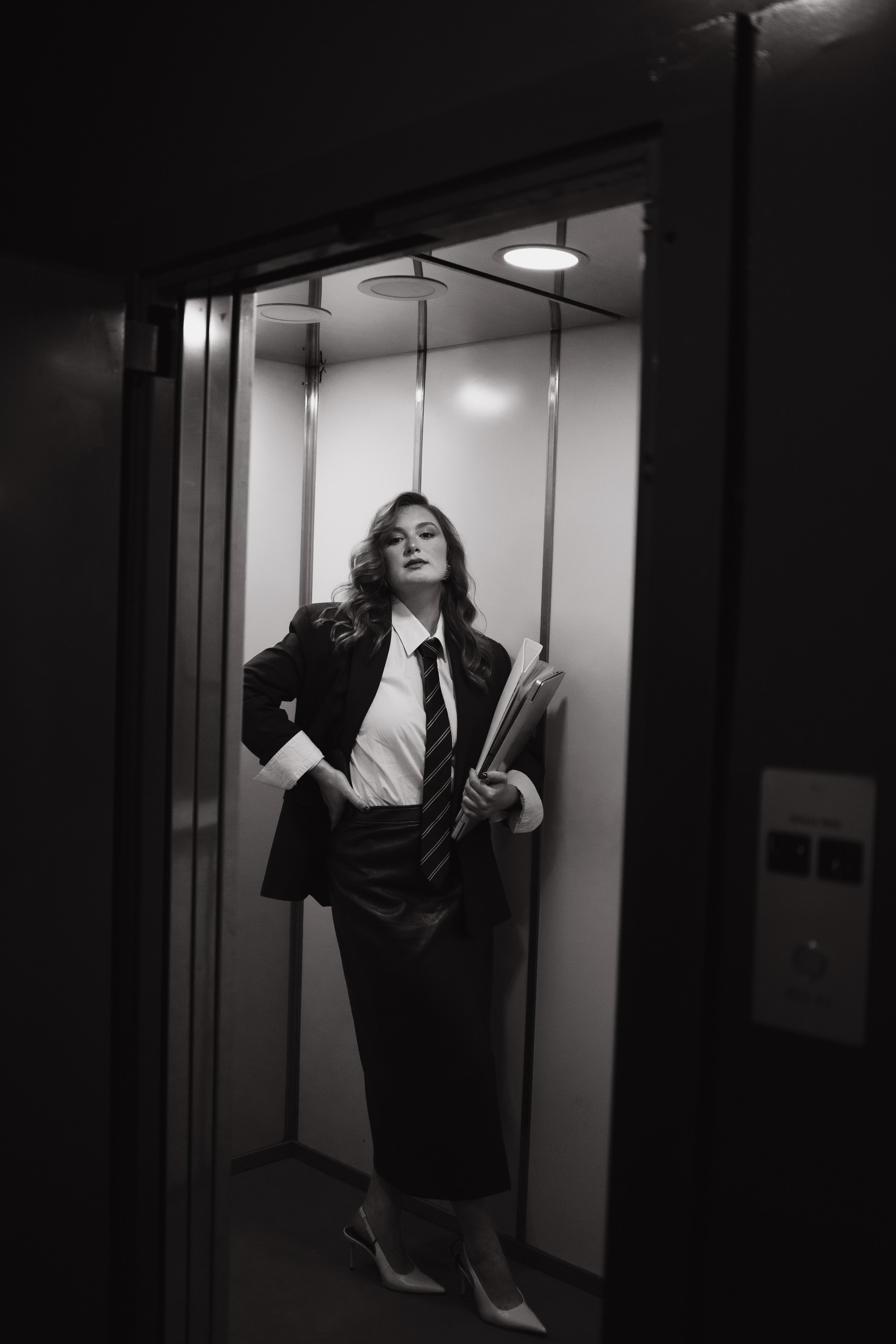 A woman with wavy hair, dressed in a suit with a tie, standing in an elevator and holding papers, seen through the elevator mirror.
