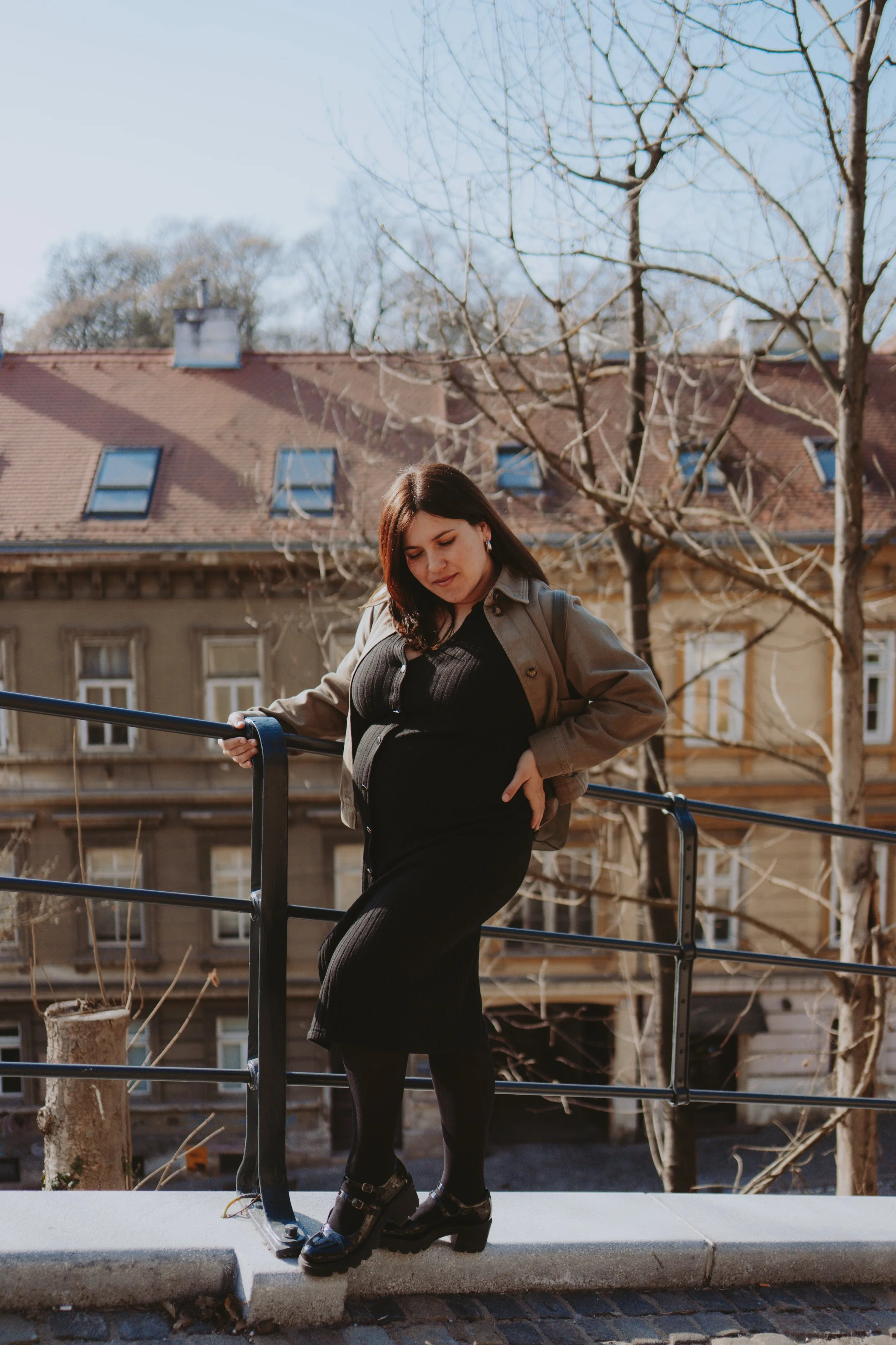 A woman with dark hair and earrings, dressed in a black dress and beige jacket, standing outdoors on a sidewalk next to a black metal railing. She is looking down and holding onto the railing with one hand, with her other hand resting on her hip. Beh