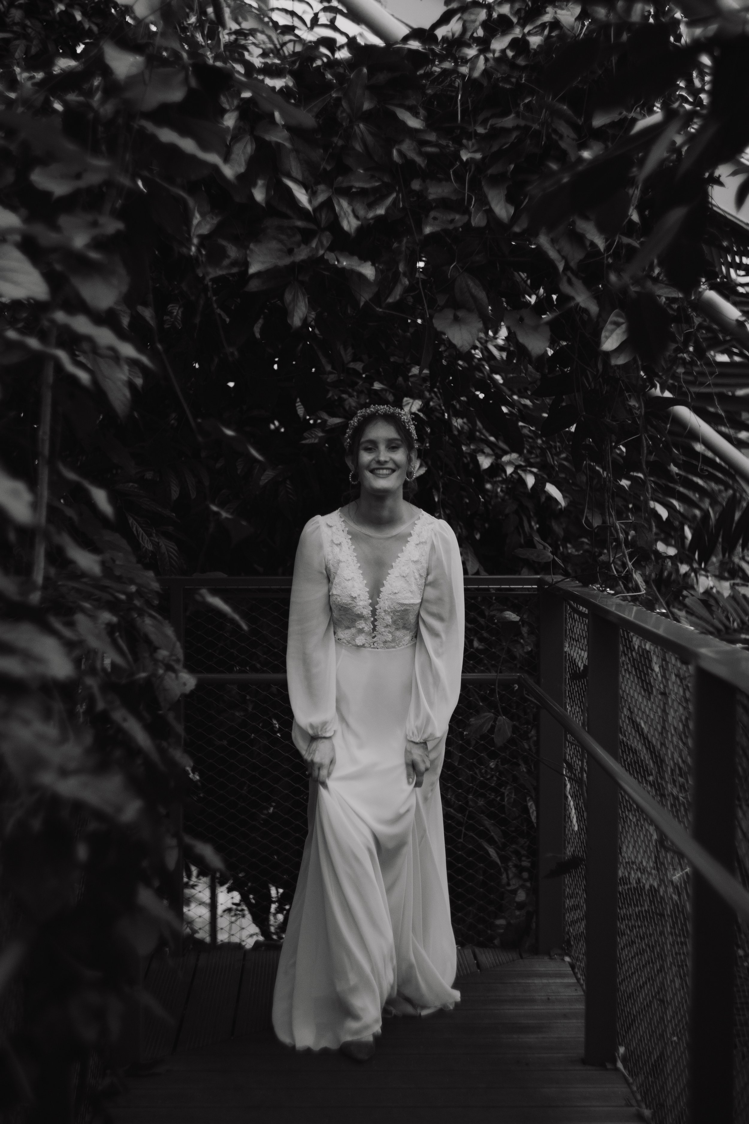A woman dressed in a long white wedding gown, smiling and posing for a photo in front of dense foliage, taken in black and white.