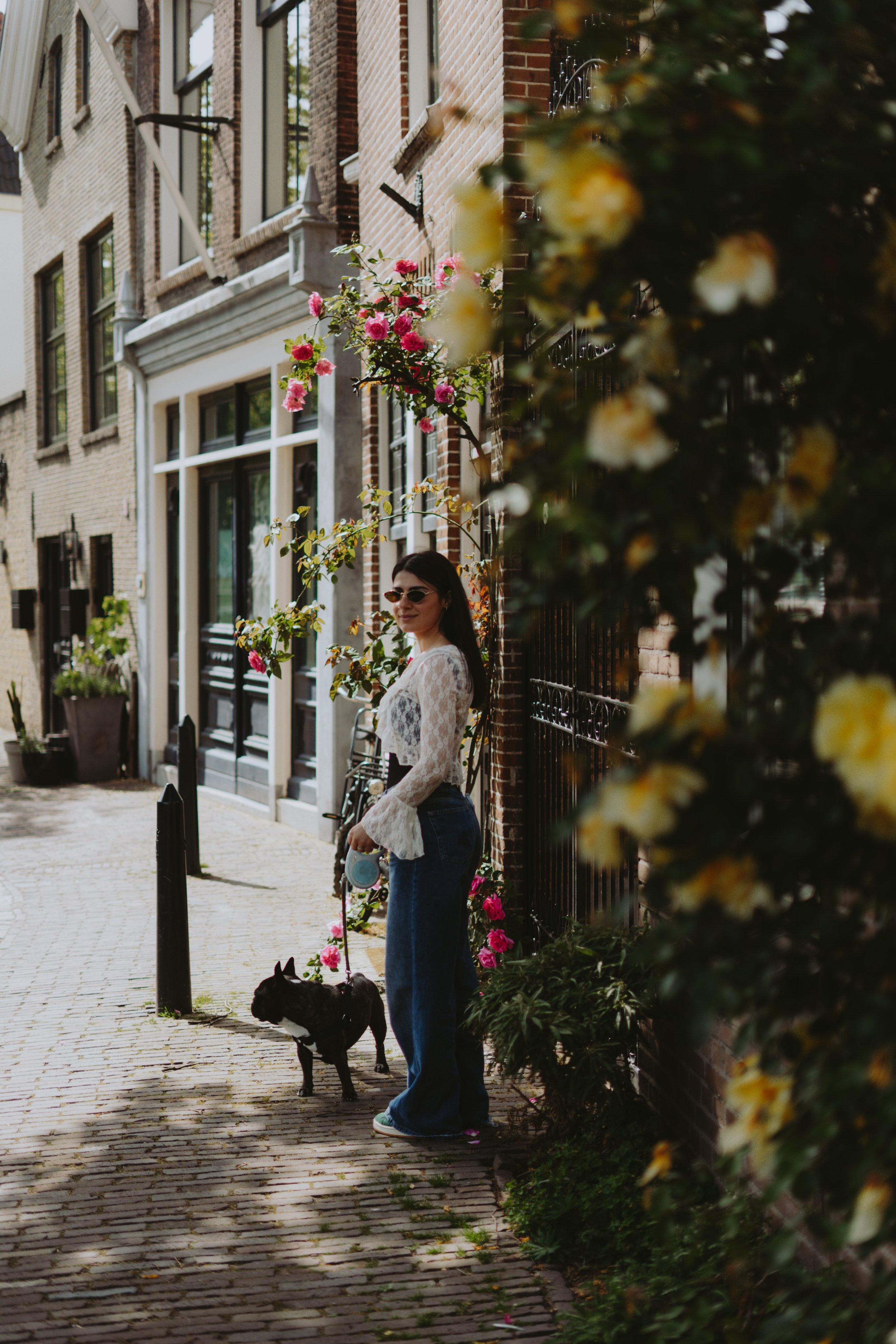 A woman wearing sunglasses, a white lace top, and blue jeans is standing on a cobblestone street with a small dog on a leash. She is smiling and positioned near a black iron fence with yellow and pink flowers in front of her. In the background, there