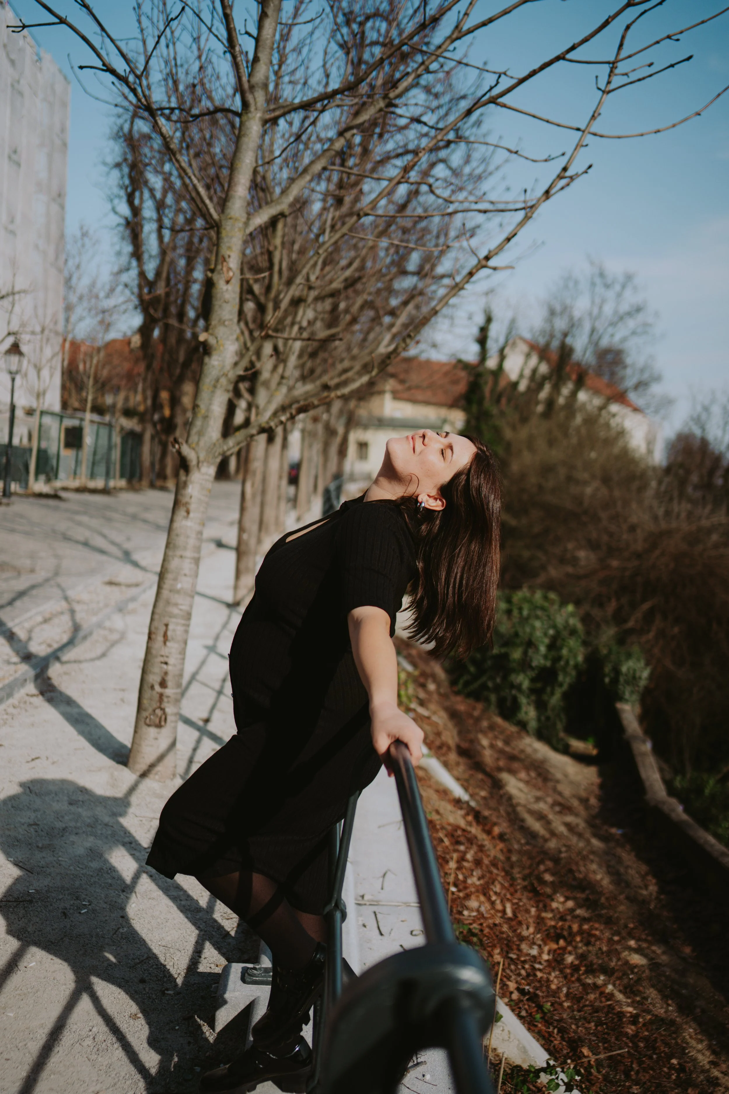 A woman with long dark hair and a black dress is taking a selfie outdoors on a sunny day, standing on a paved walkway next to a leafless tree, with houses and trees in the background.
