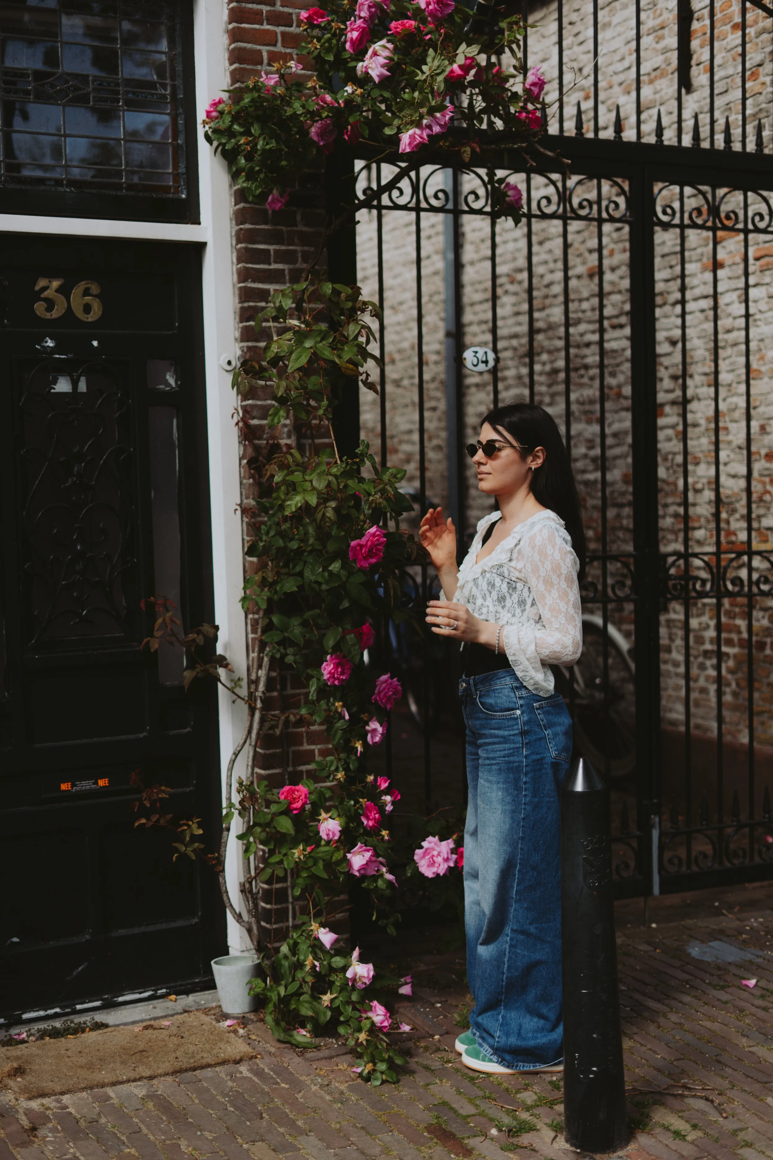 A woman wearing sunglasses, a white lace top, and wide-leg jeans standing on a cobblestone street near a black metal gate adorned with pink flowering plants. A house number 36 is visible on the door, and there is a small white and black security came