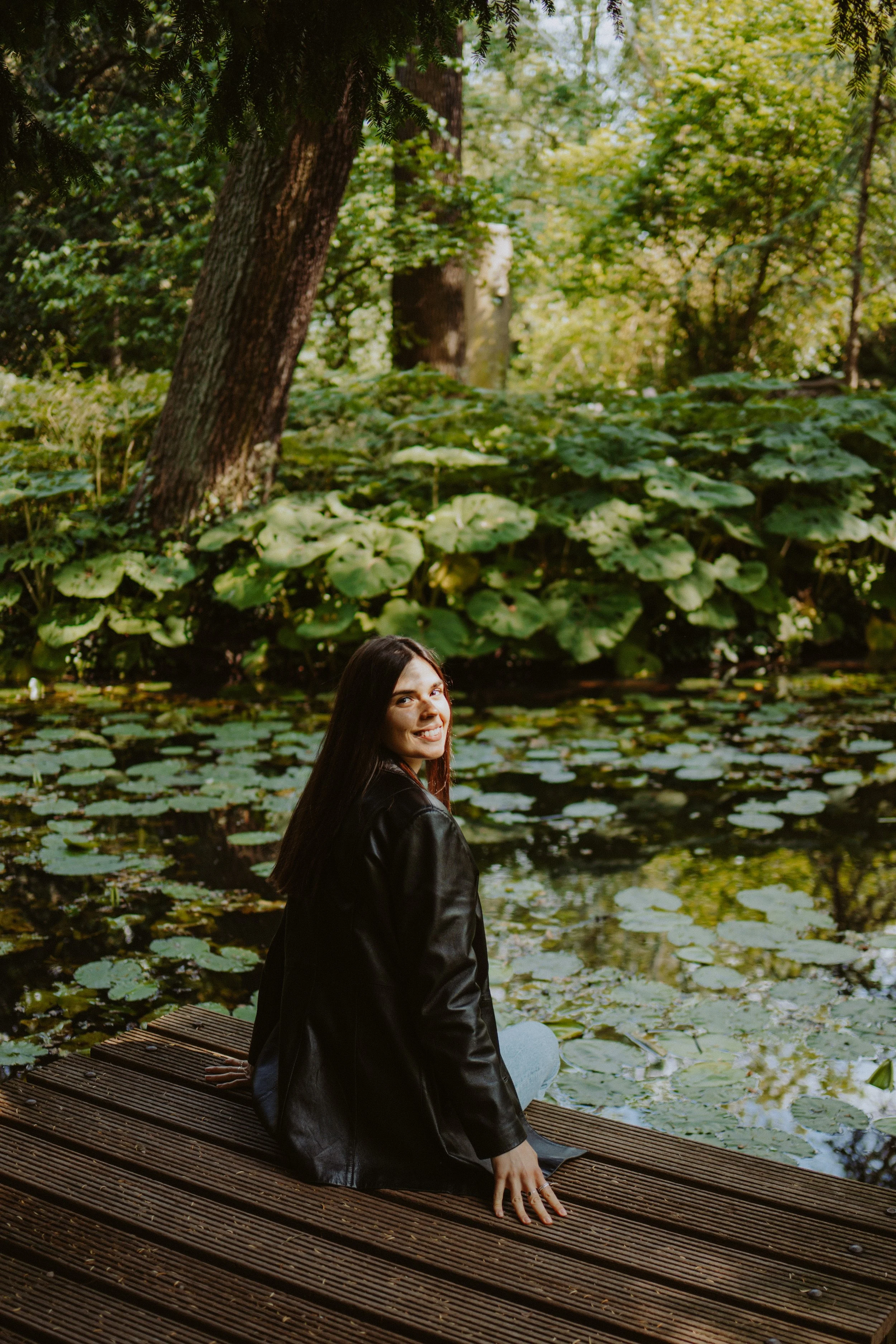 A young woman with long dark hair, wearing a black leather jacket, sitting on a wooden dock by a pond with lily pads. She is smiling and looking back over her shoulder. The background features dense greenery with trees and large leafy plants.