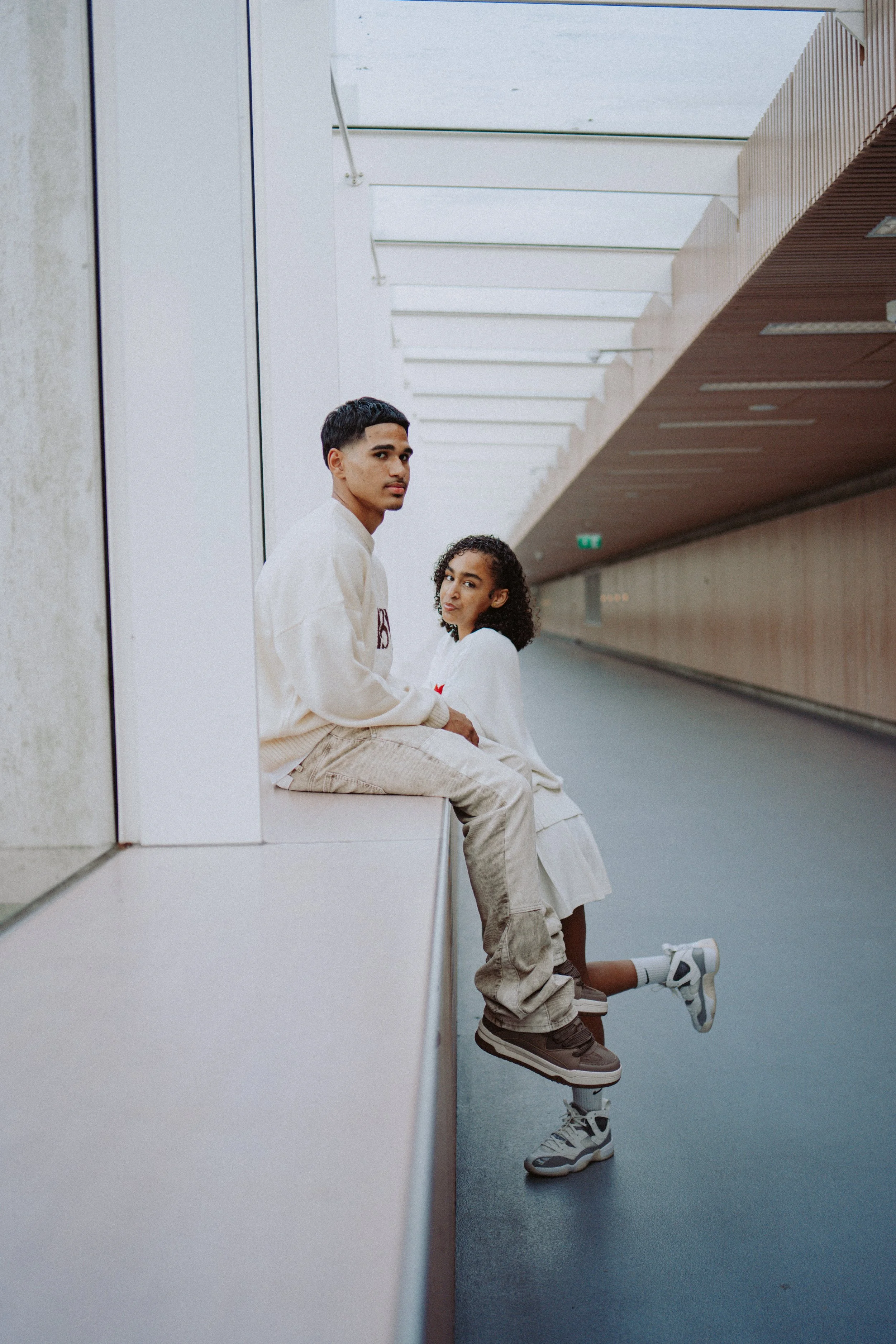 A young man and woman sitting on a ledge in a modern, architecturally designed corridor with a high ceiling and large windows, both in casual light-colored clothing.