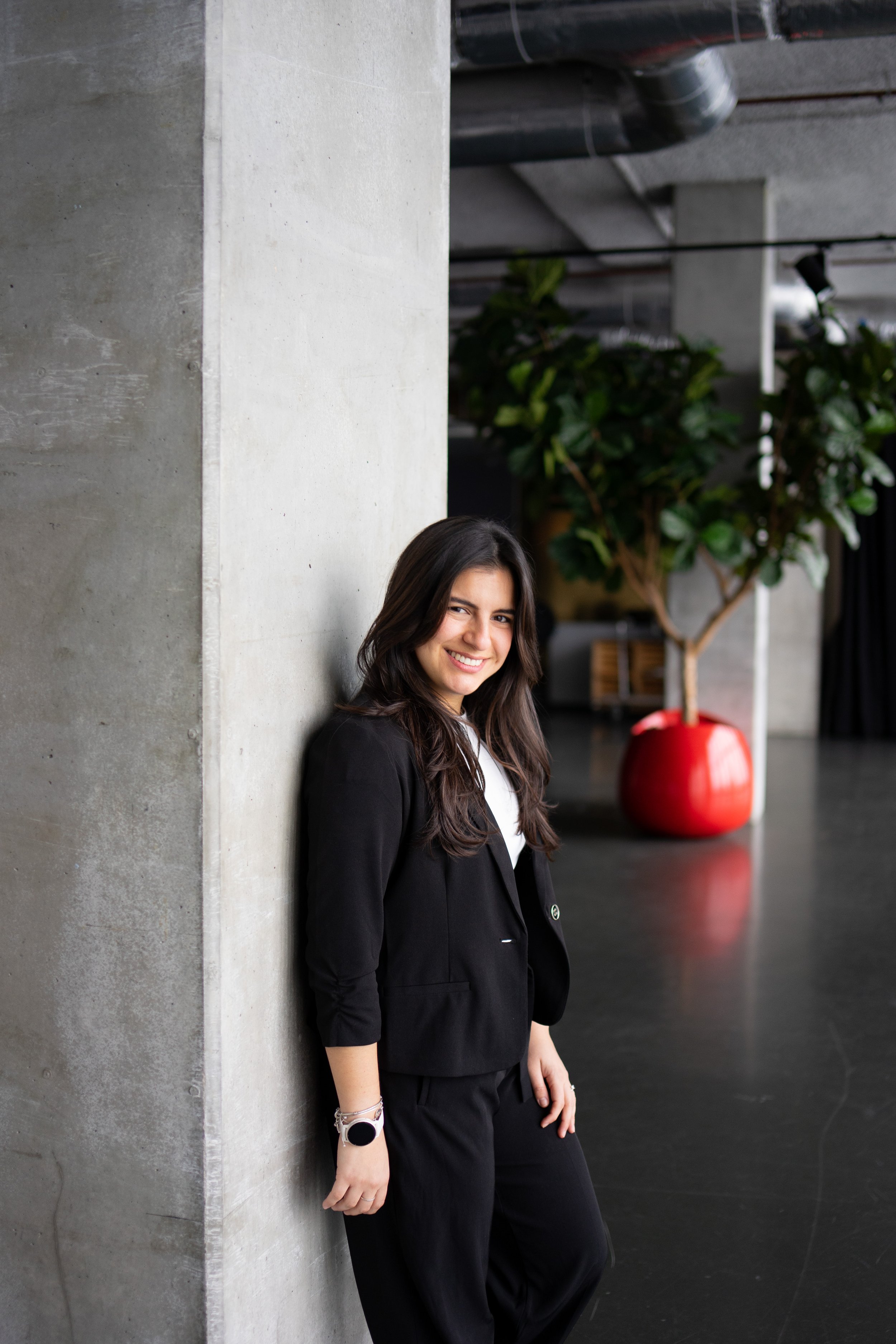A young woman with long dark hair is standing and smiling as she leans against a gray concrete wall in an indoor space with modern decor and a large potted plant in the background.