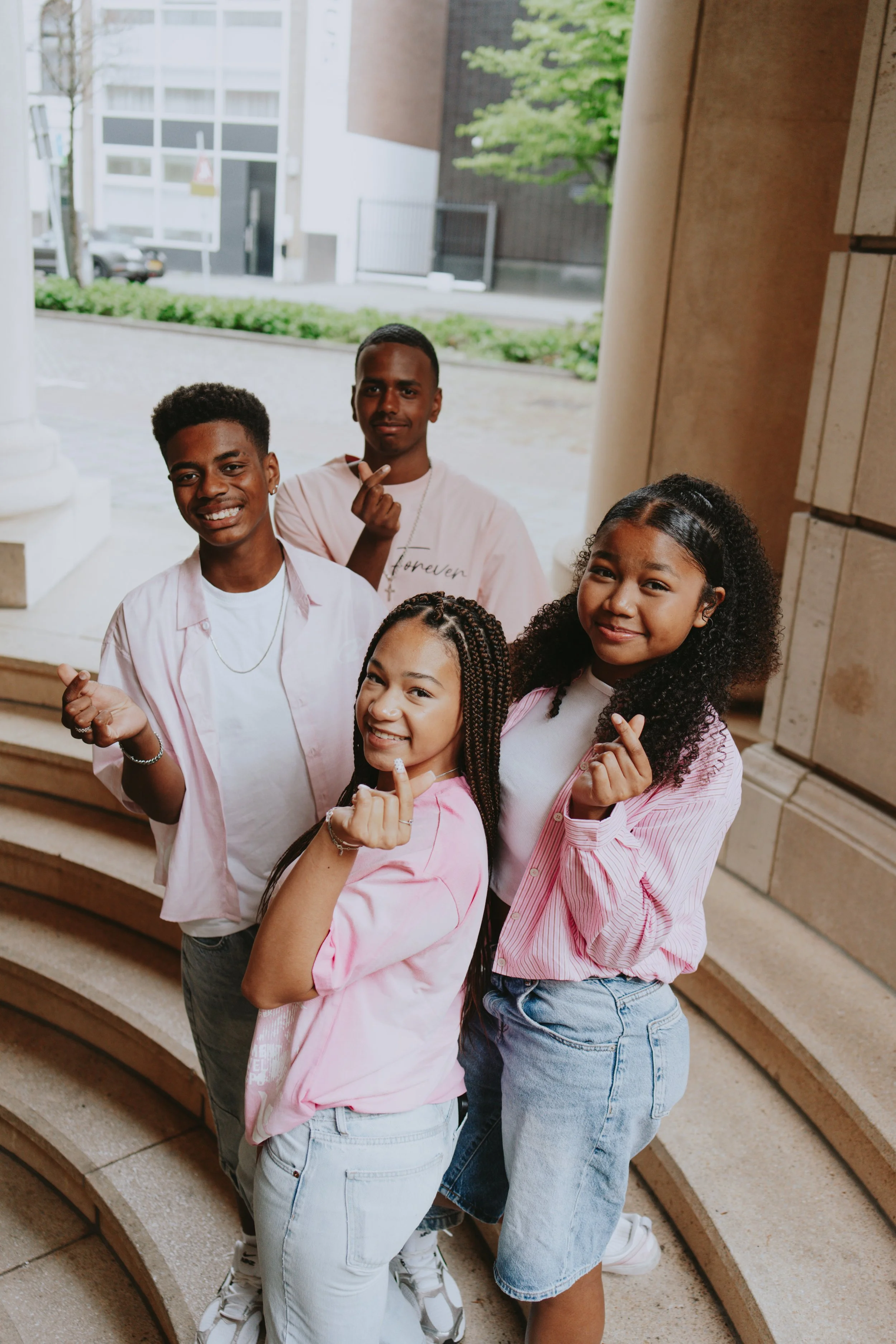 Group of five smiling young people standing on steps outside near a building, making hand gestures and wearing casual clothes.