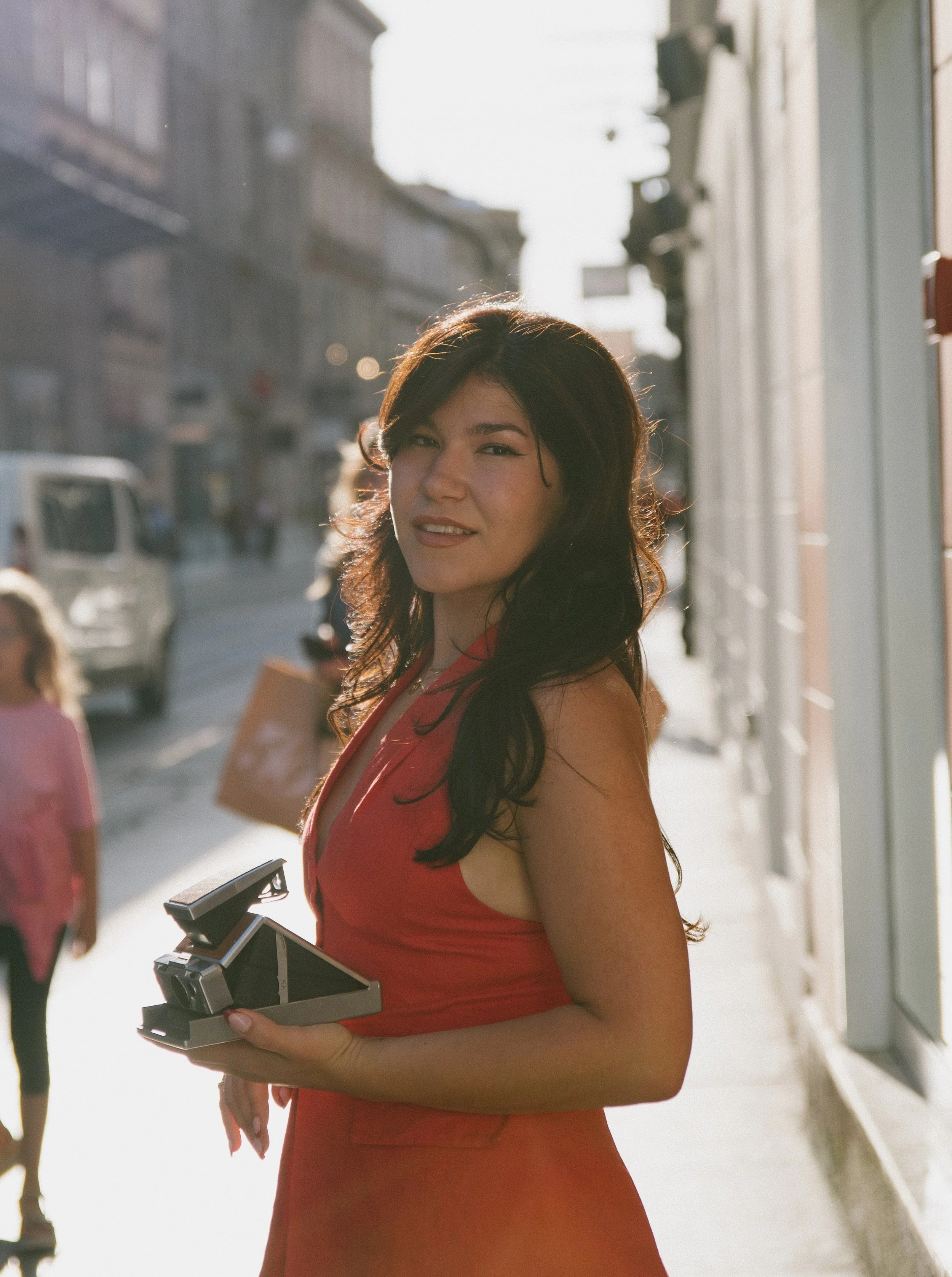A woman in a red dress holding a vintage film camera in an outdoor street setting, lit by sunlight.
