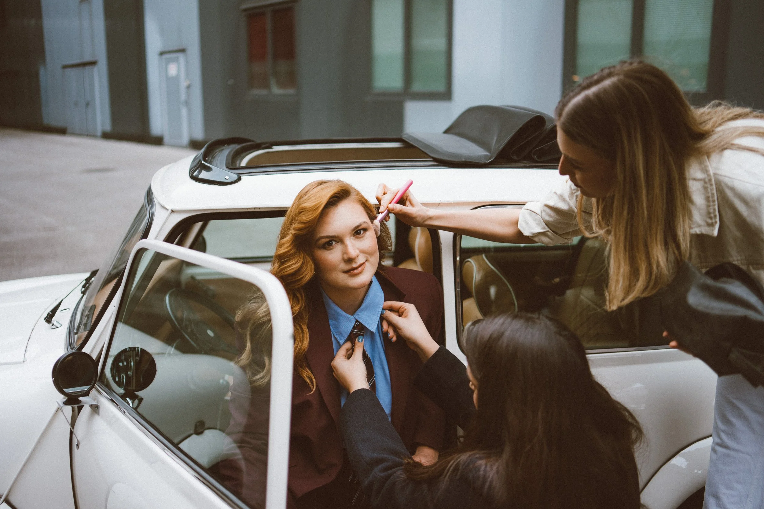Branding photoshoot of a woman, capturing behind the scene with a professional MUA applying makeup or touching her face with a makeup brush. Several people are surrounding the car, assisting with her appearance.