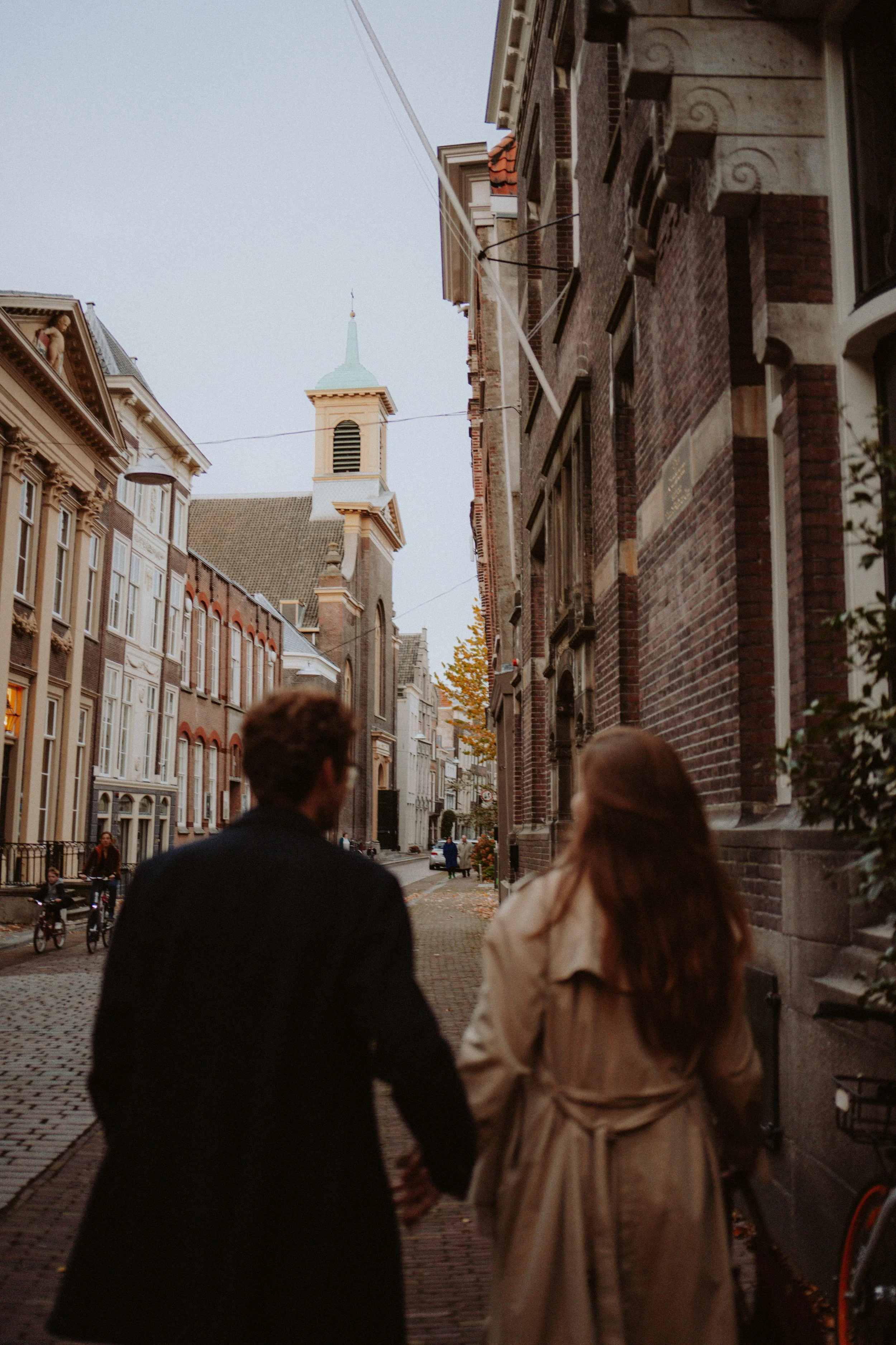 A couple walking hand in hand on a cobblestone street in a European city, with historic brick and stone buildings lining the street, and a church steeple visible in the background.