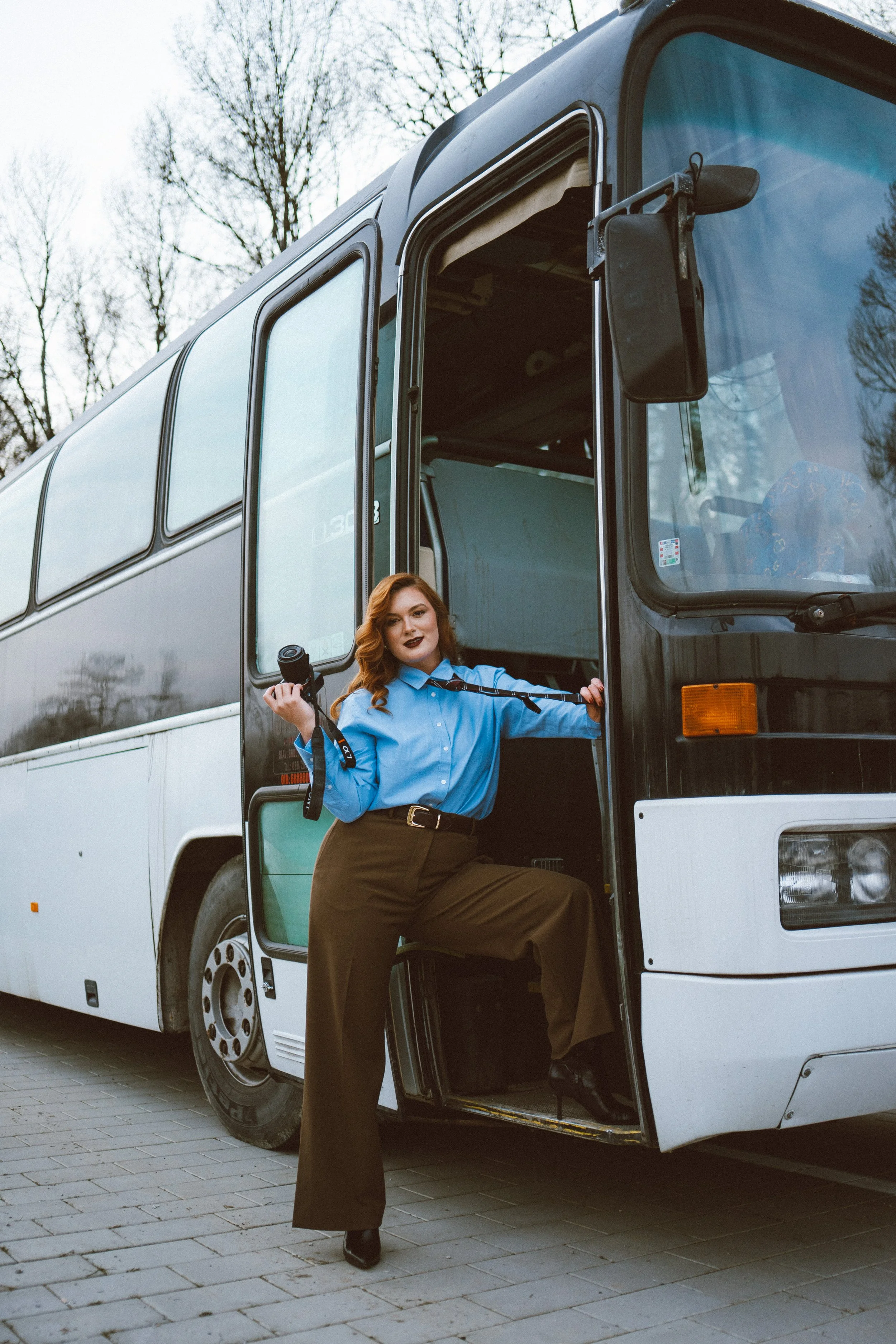 Editorial photoshoot of a woman in a blue shirt and brown pants standing at the door of a large white bus, holding a camera strap, with trees and an overcast sky in the background.