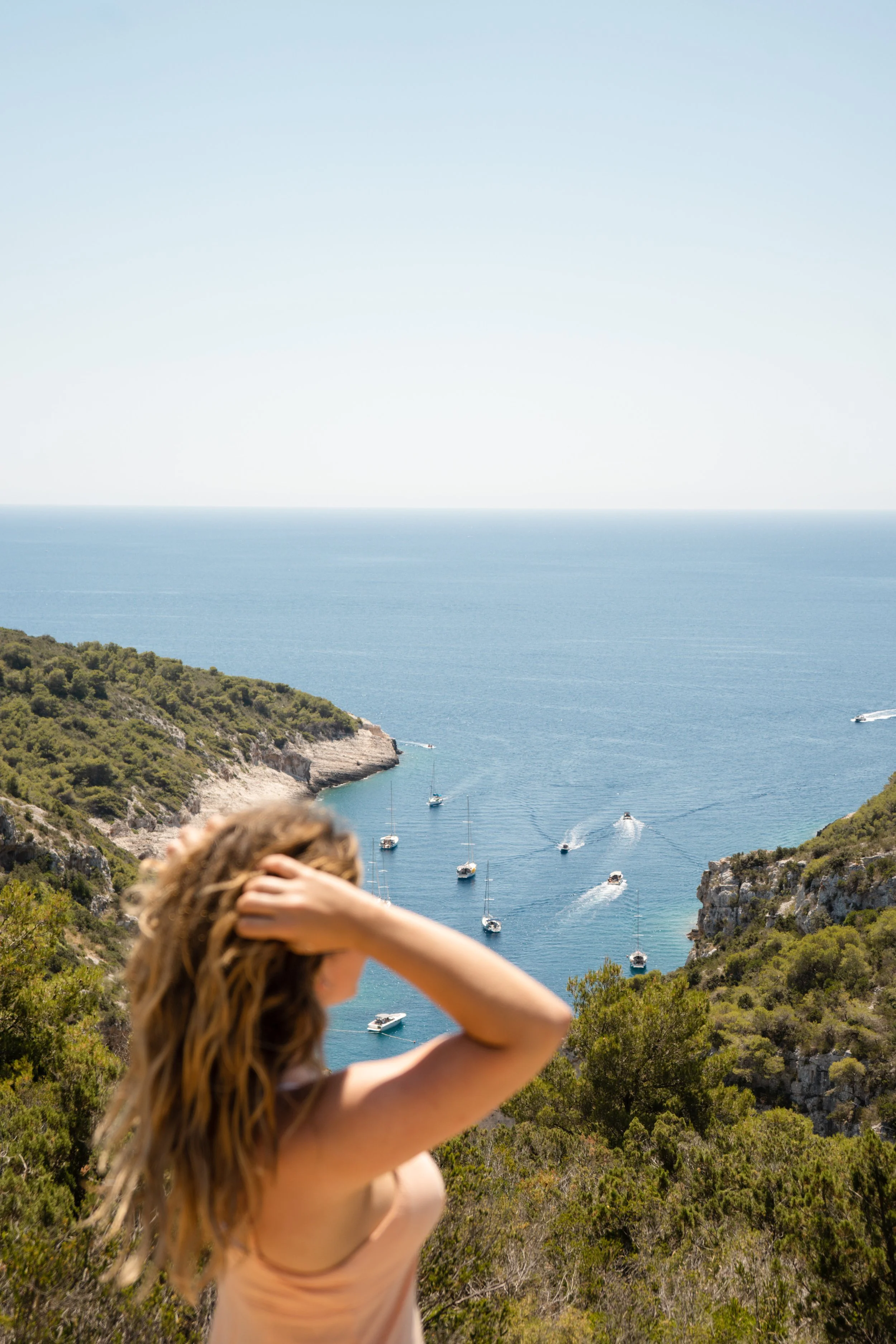 A woman with long wavy hair stands with her back to the camera on a hillside overlooking a blue ocean with several sailboats, wearing a sleeveless top and holding her hair with one hand.