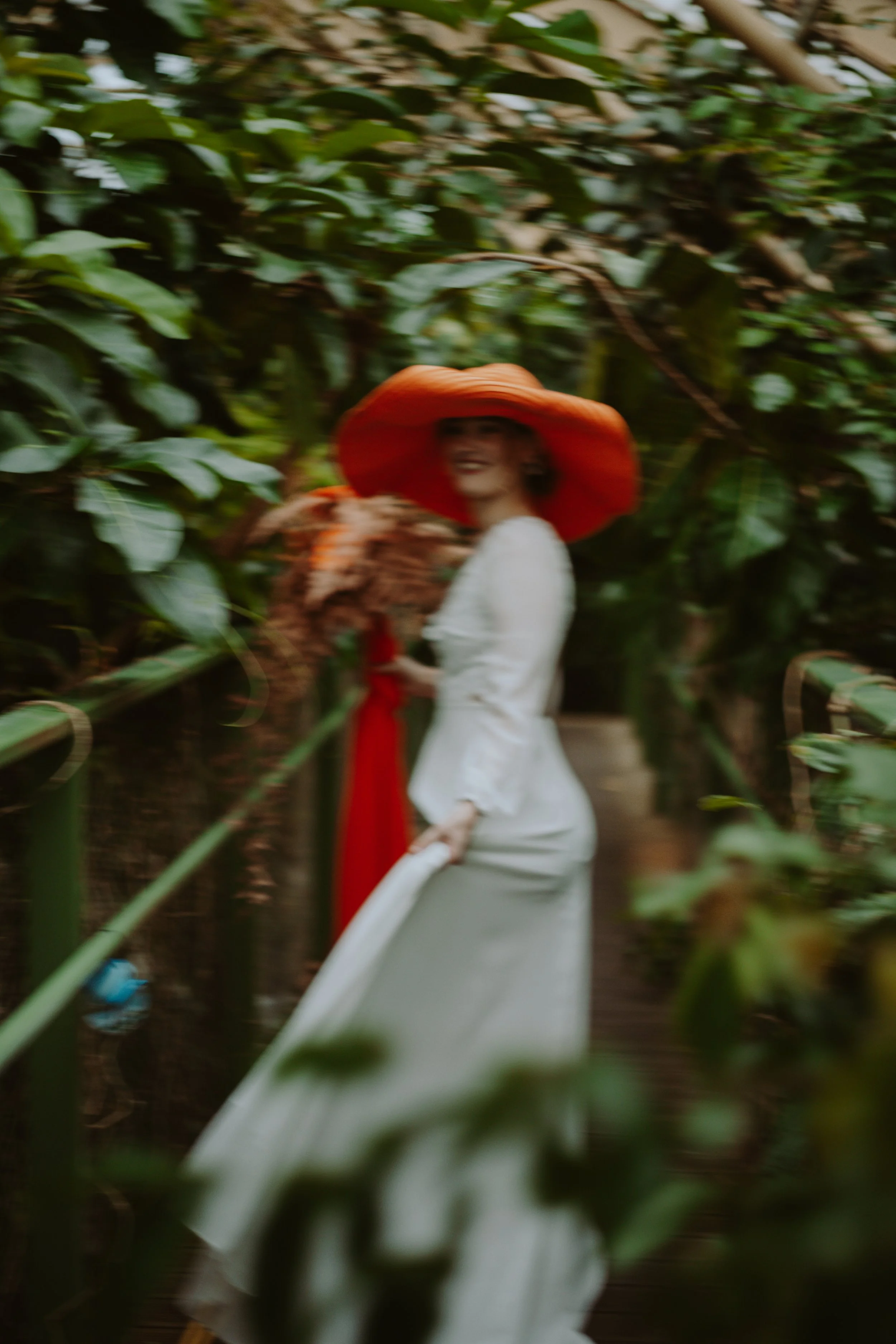 A woman in a white dress and large orange hat standing among green plants, smiling and holding a chicken.