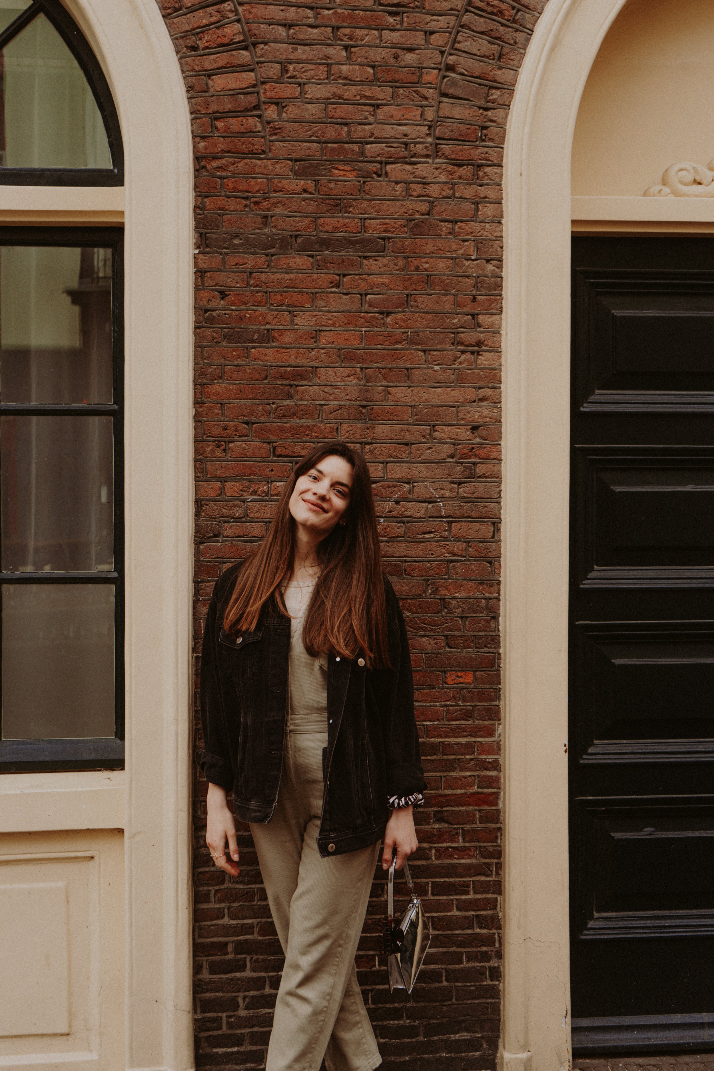 Young woman with long brown hair smiling, standing outside against a brick wall between two doors, one glass-paneled and one black, in an urban setting.