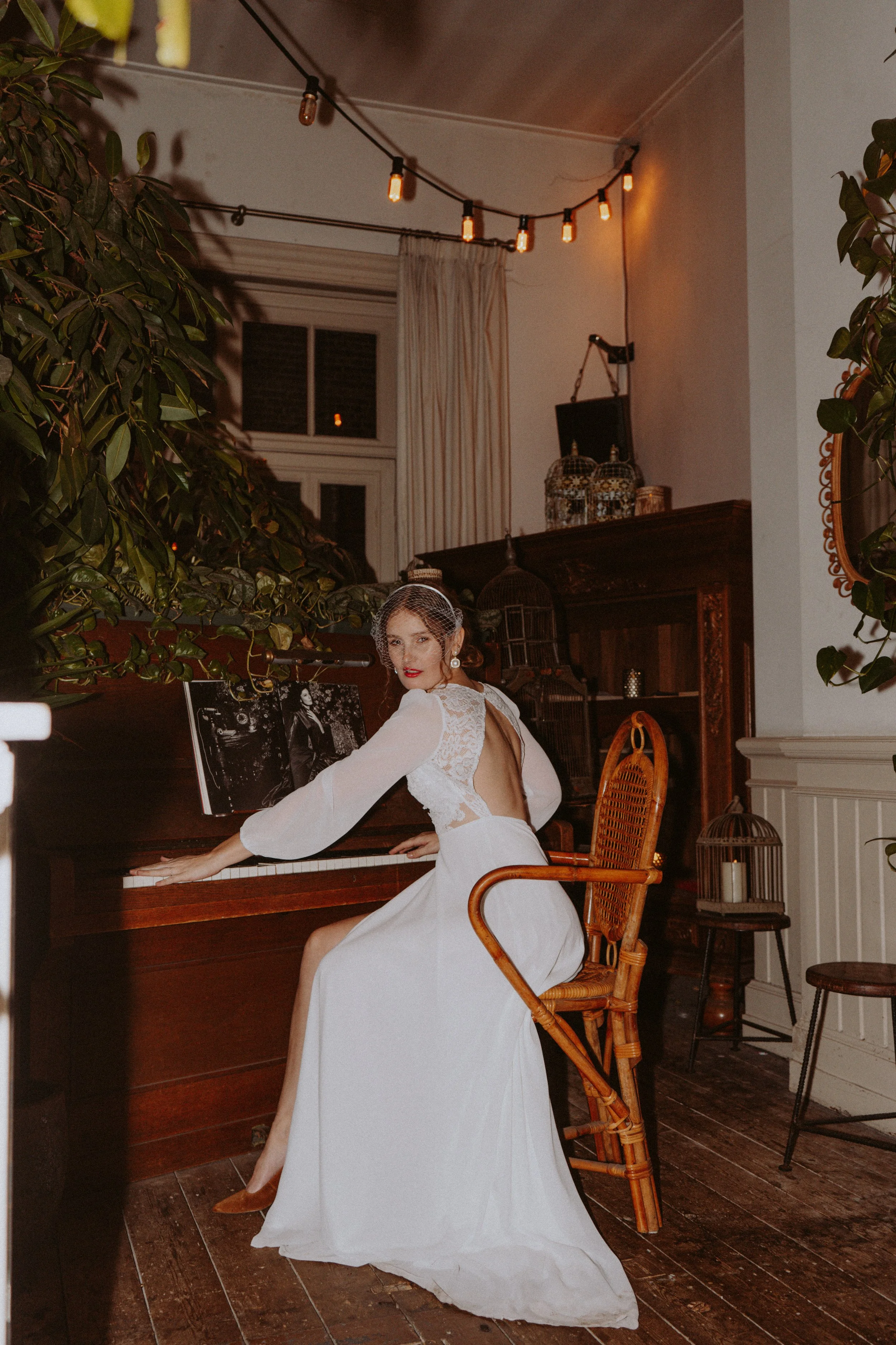 A woman in a white dress with lace details is sitting at a piano in a cozy, decorated room. She is wearing vintage-style earrings, a small veil, and makeup with red lipstick. The room has warm lighting, potted plants, framed photos on the piano, a bi