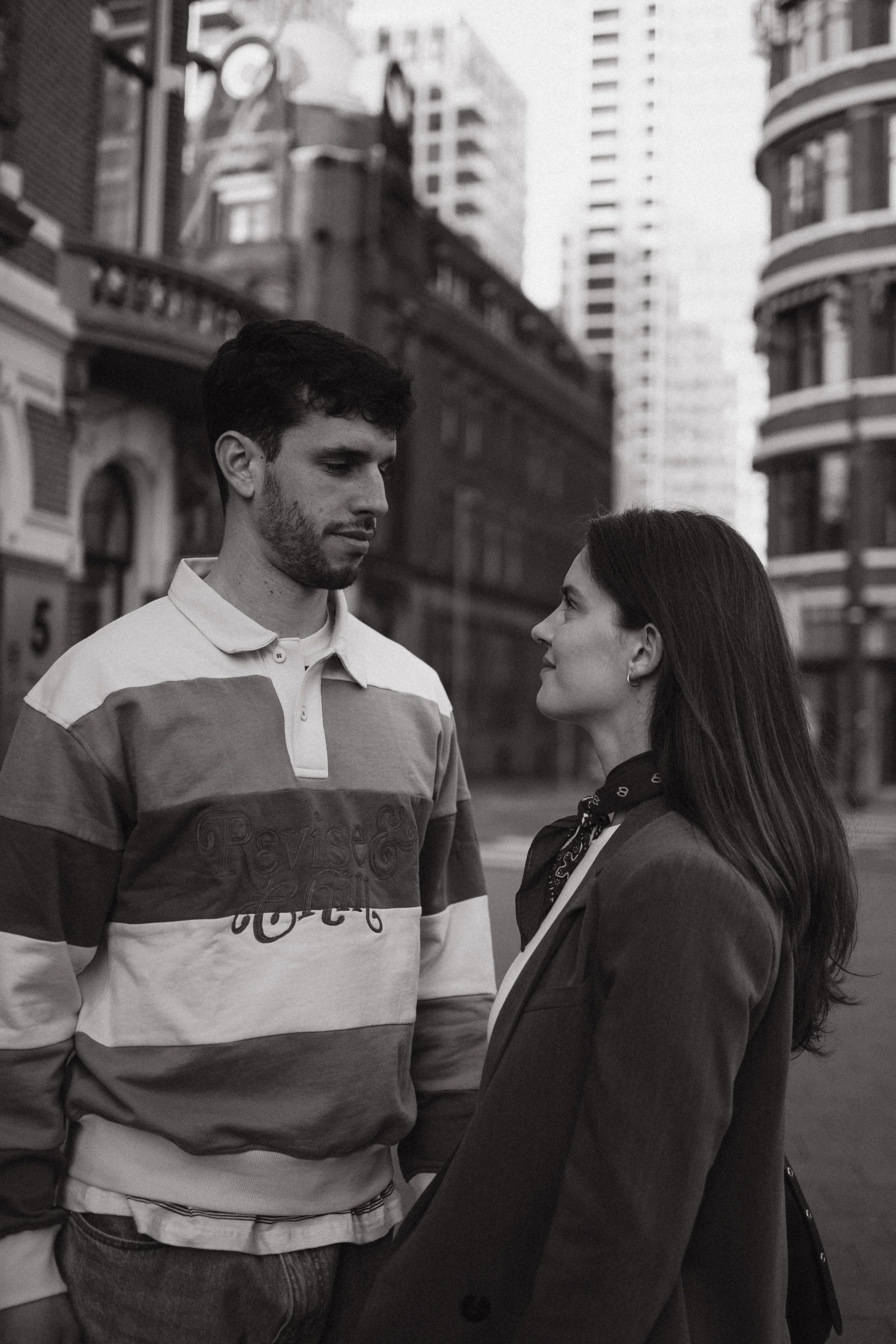 A man and woman face each other on a city street with tall buildings in the background.