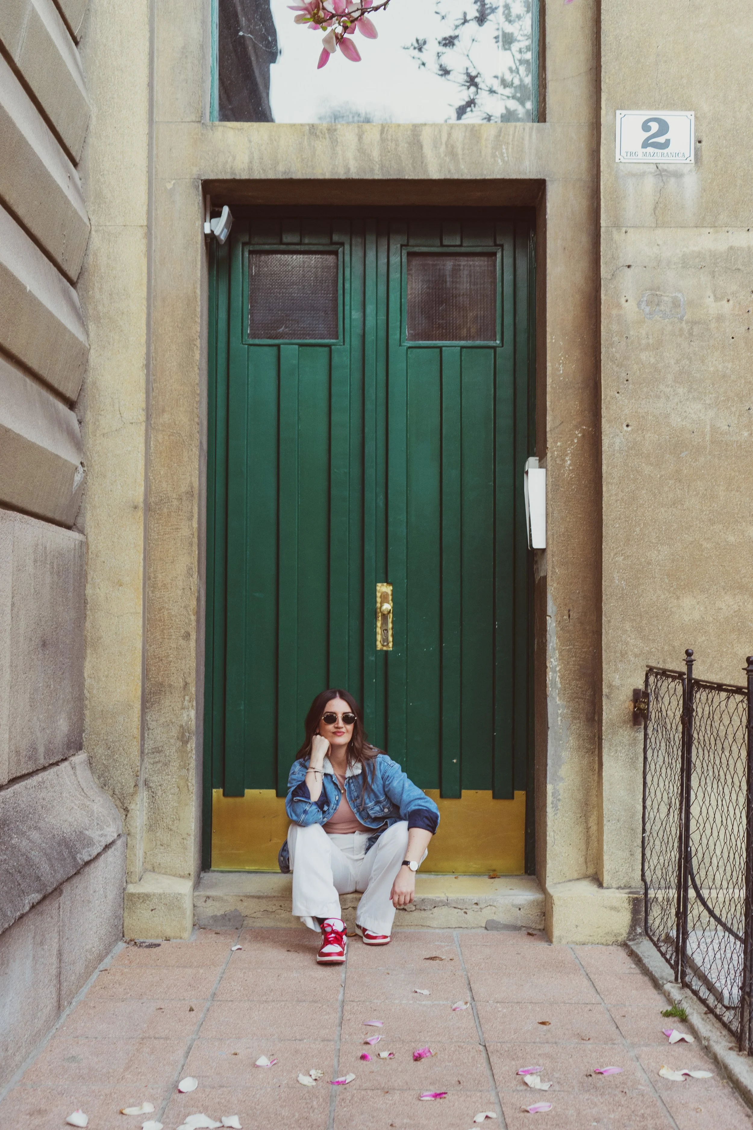 A woman sitting on the front steps of a building, wearing sunglasses, a denim jacket, and white pants, with pink sneakers. The building has a large green door, a small window above, and flower petals scattered on the ground.