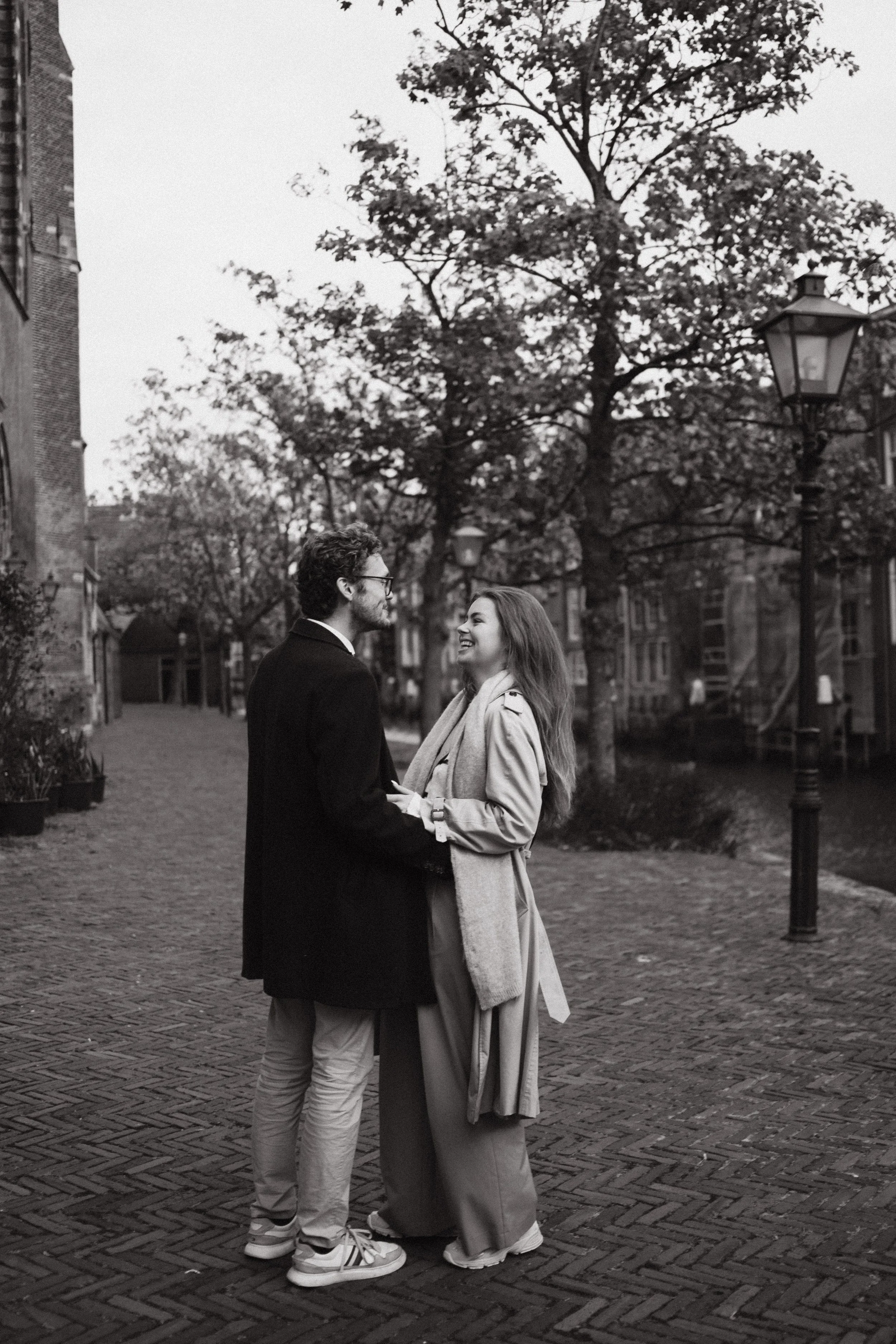 Black and white photography of a couple in Dordrecht standing on a cobblestone street, holding hands and looking at each other, with trees and buildings in the background.