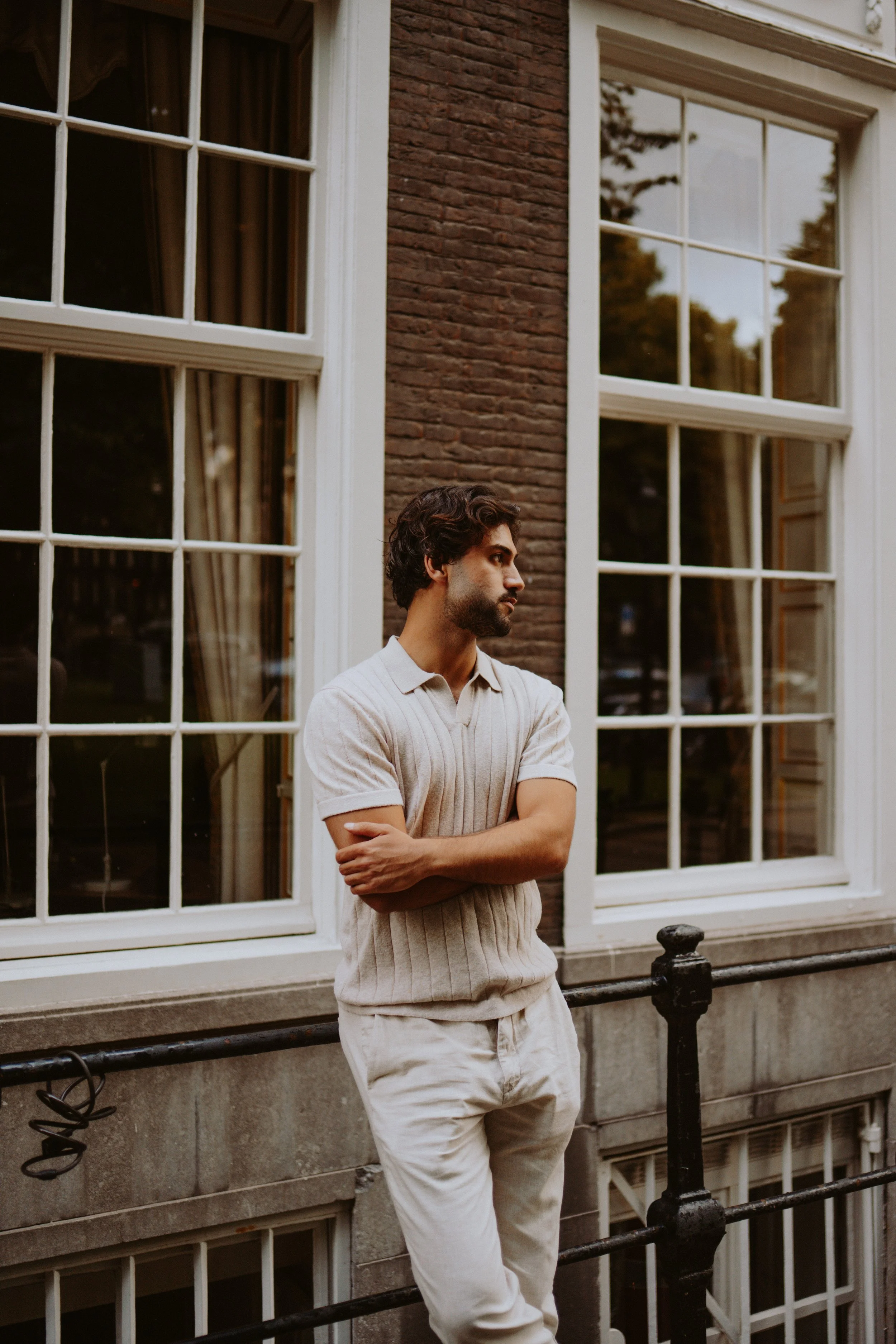 A man with dark curly hair and a beard, wearing a beige short-sleeve sweater and light-colored pants, stands outside in front of a large window with white framing on a brick building, leaning against a black metal railing, looking to the side with ar