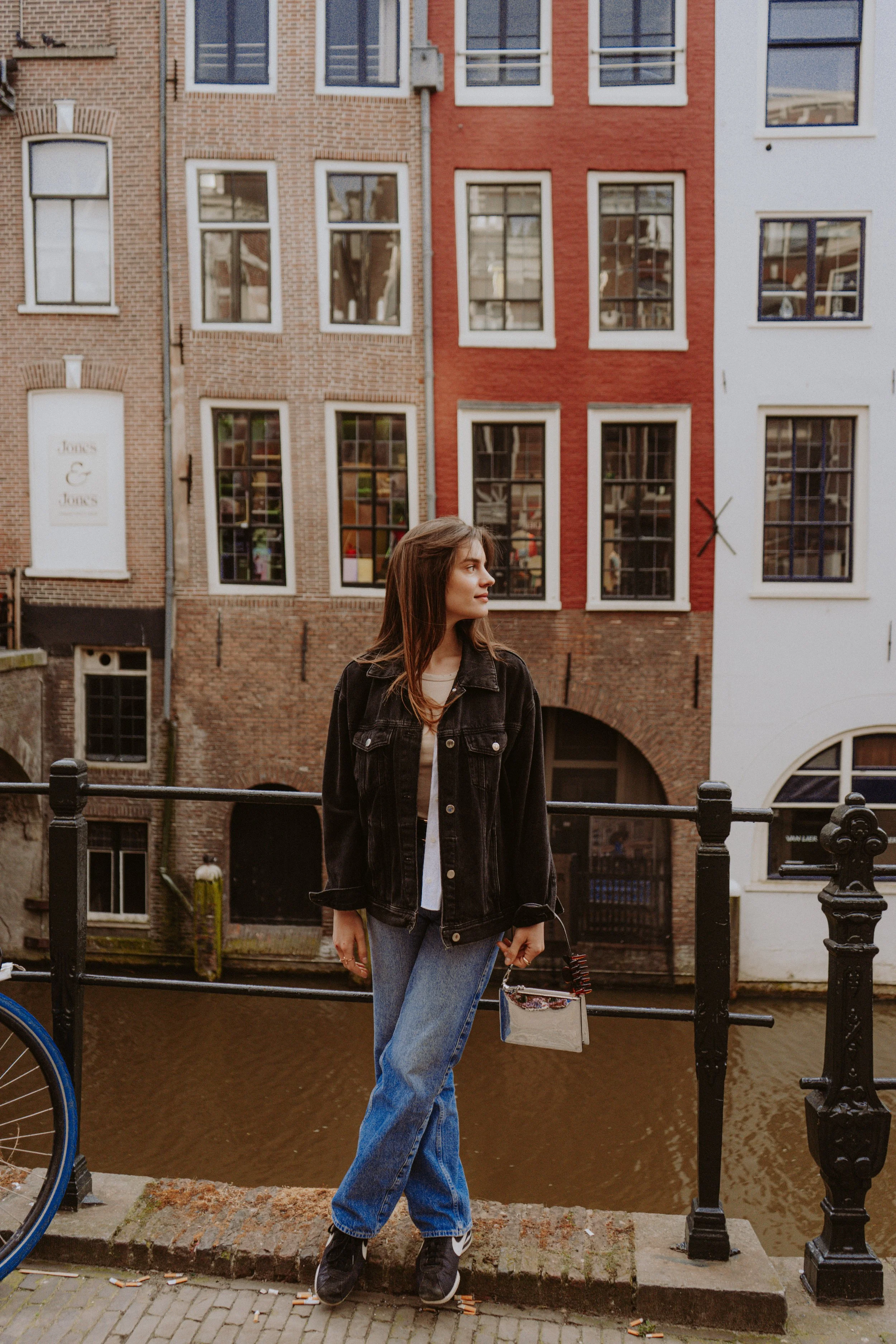 A young woman with brown hair wearing a black denim jacket and blue jeans standing on a bridge over a canal, holding a small handbag. In the background are brick buildings with multiple windows and a canal. A bicycle wheel is partly visible on the le