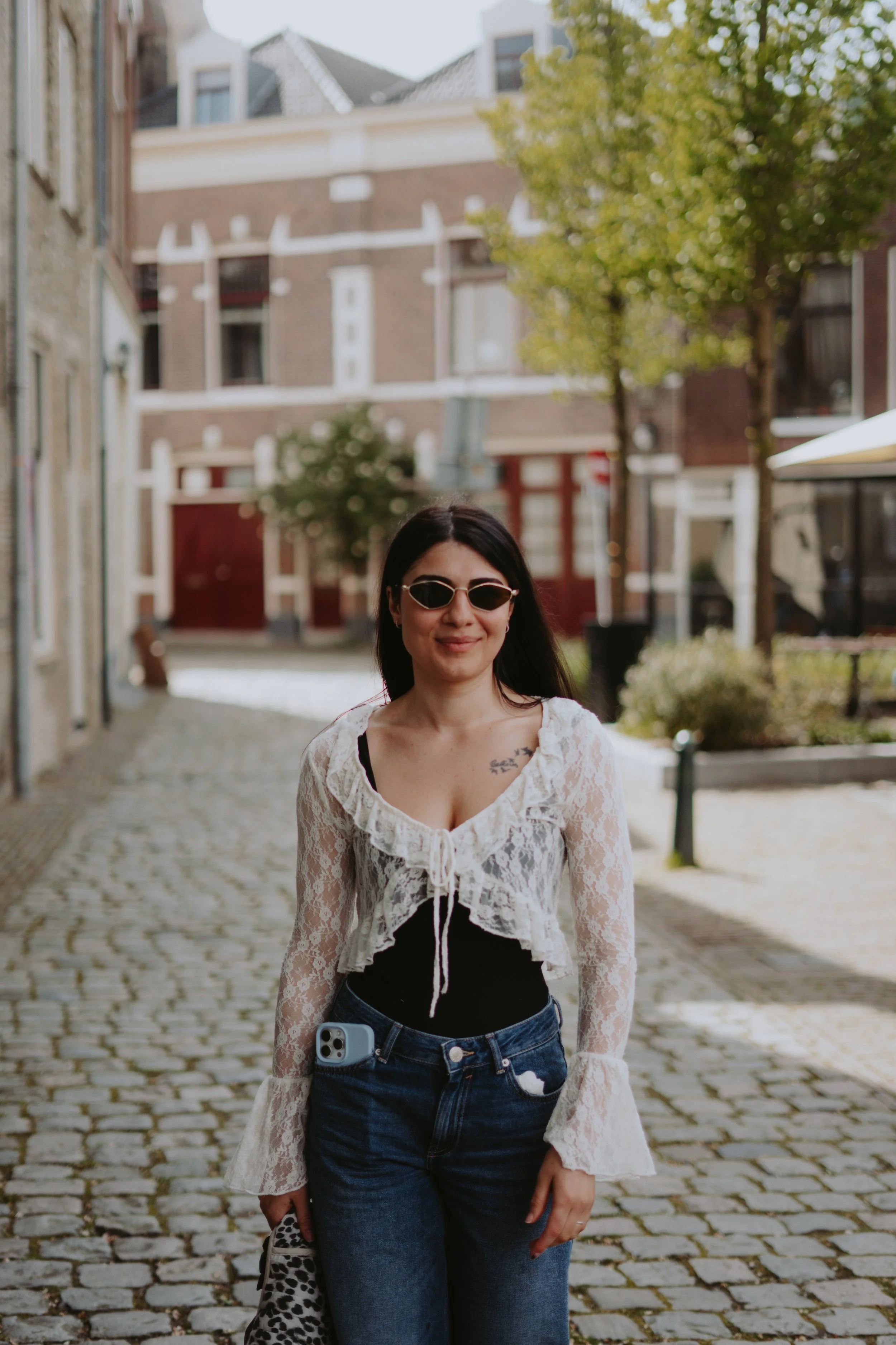 A woman with black hair wearing sunglasses, a white lace top, and jeans is standing on a cobblestone street, smiling with a tattoo on her chest, holding a phone with a leopard print case, and a phone attached to her jeans.