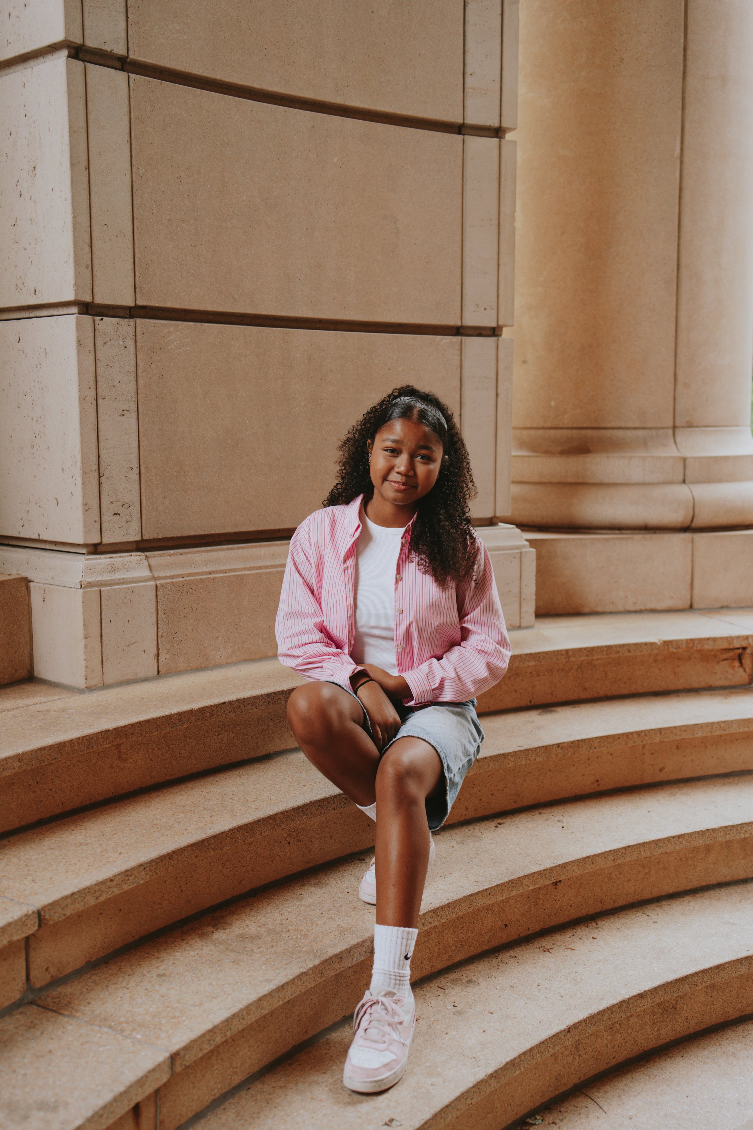 A young girl with curly hair sitting on steps outside a large stone building, smiling, wearing a pink and white striped shirt, denim shorts, white Nike socks, and light-colored sneakers.